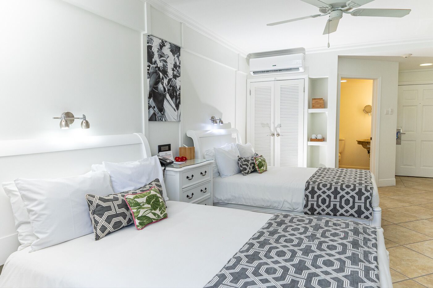 A hotel room with two beds, a ceiling fan, an air conditioning unit, a bedside table with a lamp, and a wardrobe.