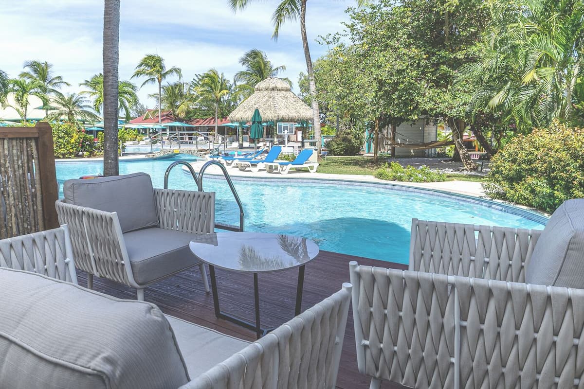 Zona junto a la piscina con sillas de descanso y una mesa, con vista a una piscina que también tiene sillas de descanso y sombrillas.