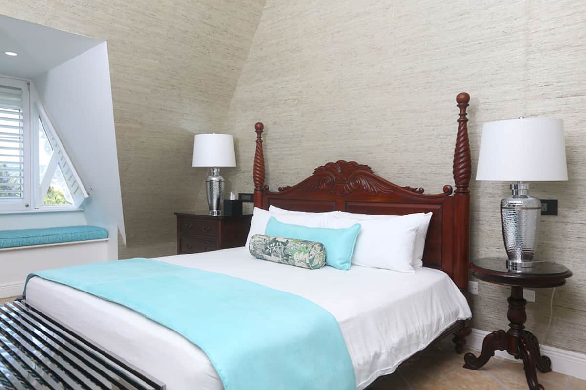 Una cama bien tendida con cabecera de madera, una manta color turquesa y una almohada estampada. Hay dos mesitas de noche con lámparas y una ventana con persianas a la izquierda.