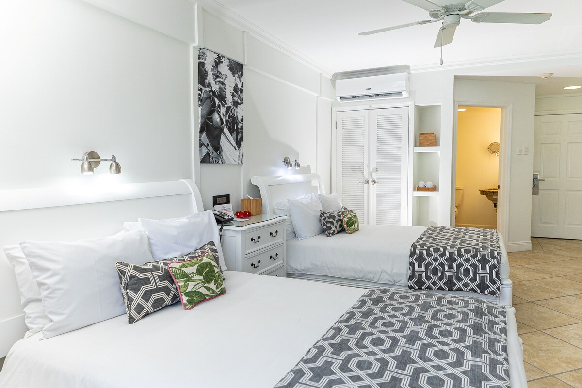 Une chambre avec deux lits, du linge de lit blanc, des coussins à motifs, une table de chevet avec une lampe, un ventilateur de plafond et un appareil de climatisation.