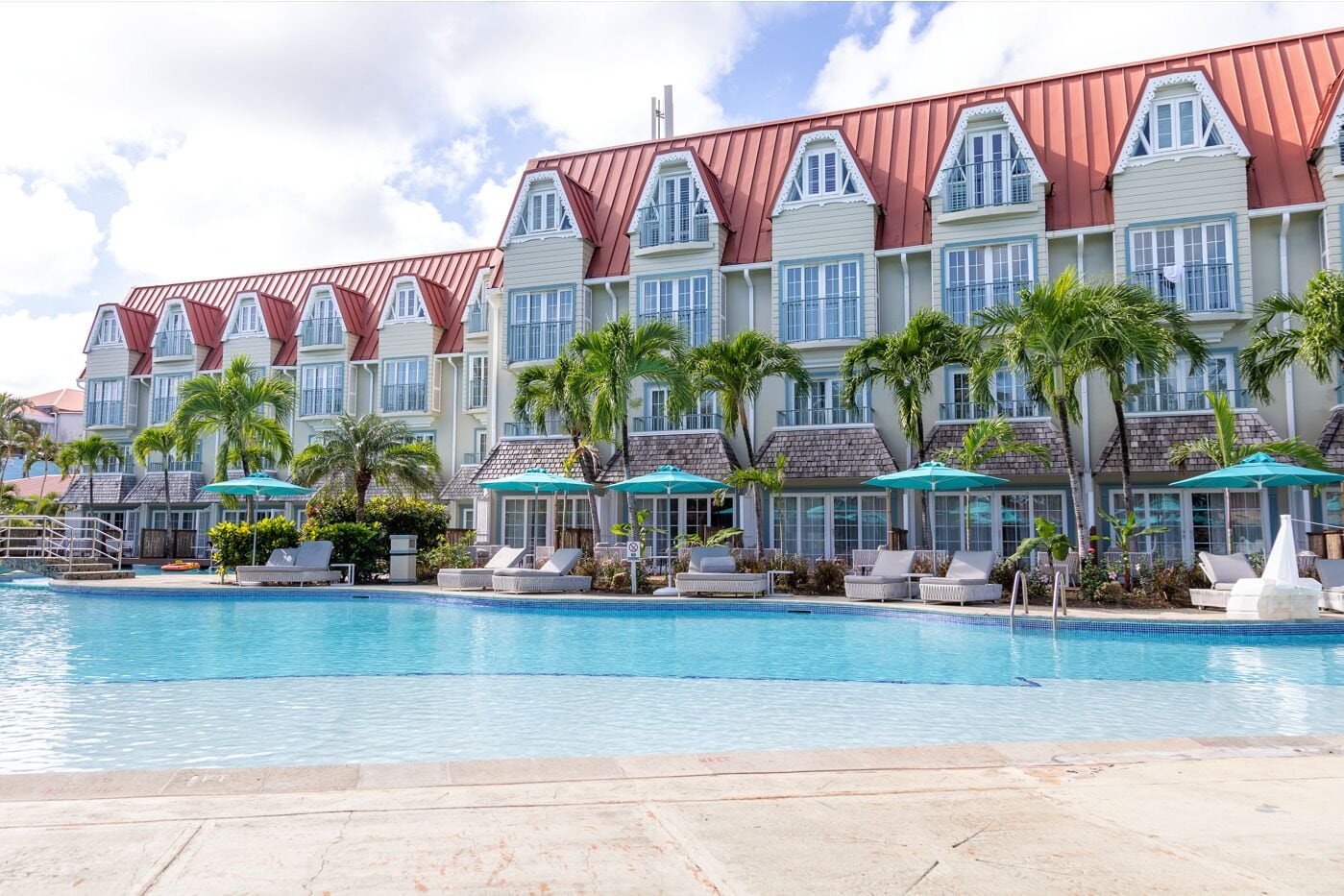 A hotel with a large pool, multiple lounge areas with umbrellas, and a row of palm trees.