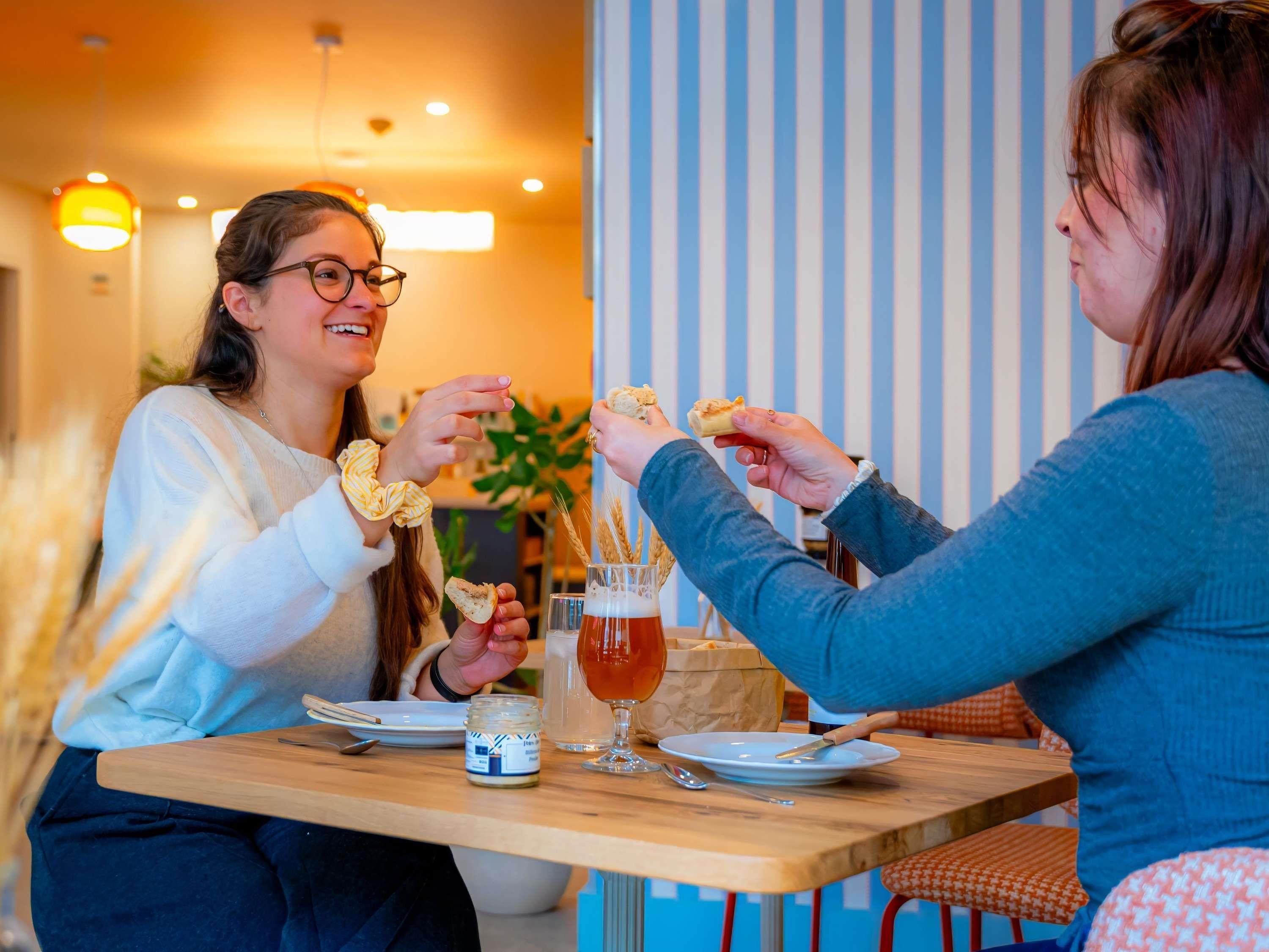 Two people sitting at a wooden table with a glass of beer and bread, in a room with striped walls and a plant in the background.