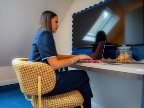 A person seated on a patterned chair at a desk with a laptop, a smartphone, and a jar of snacks. - ibis Styles Nantes Centre Place Graslin (Nantes)