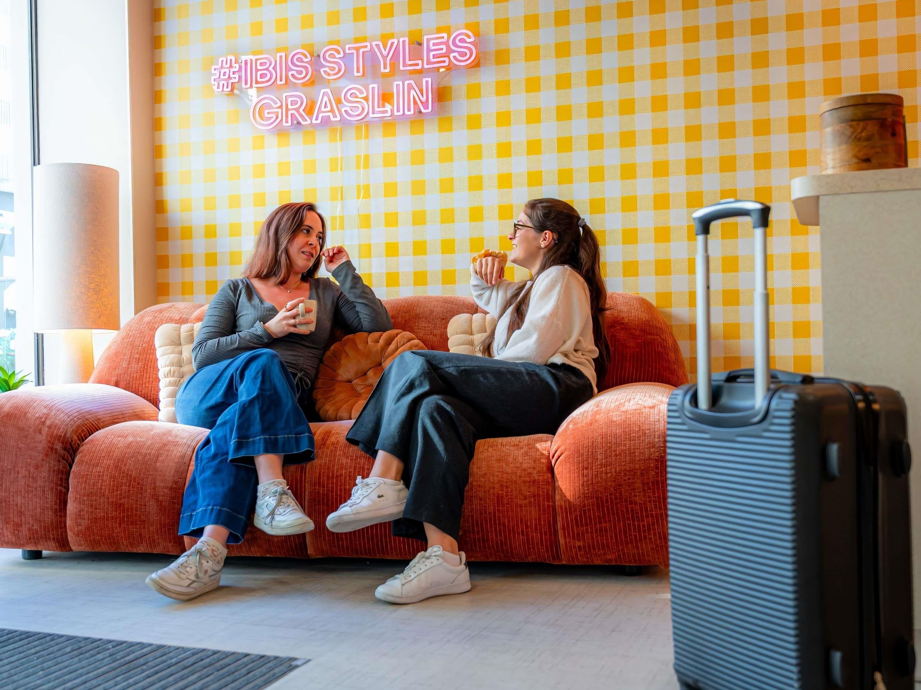 Two people sitting on a checkered orange couch with a neon sign in the background.