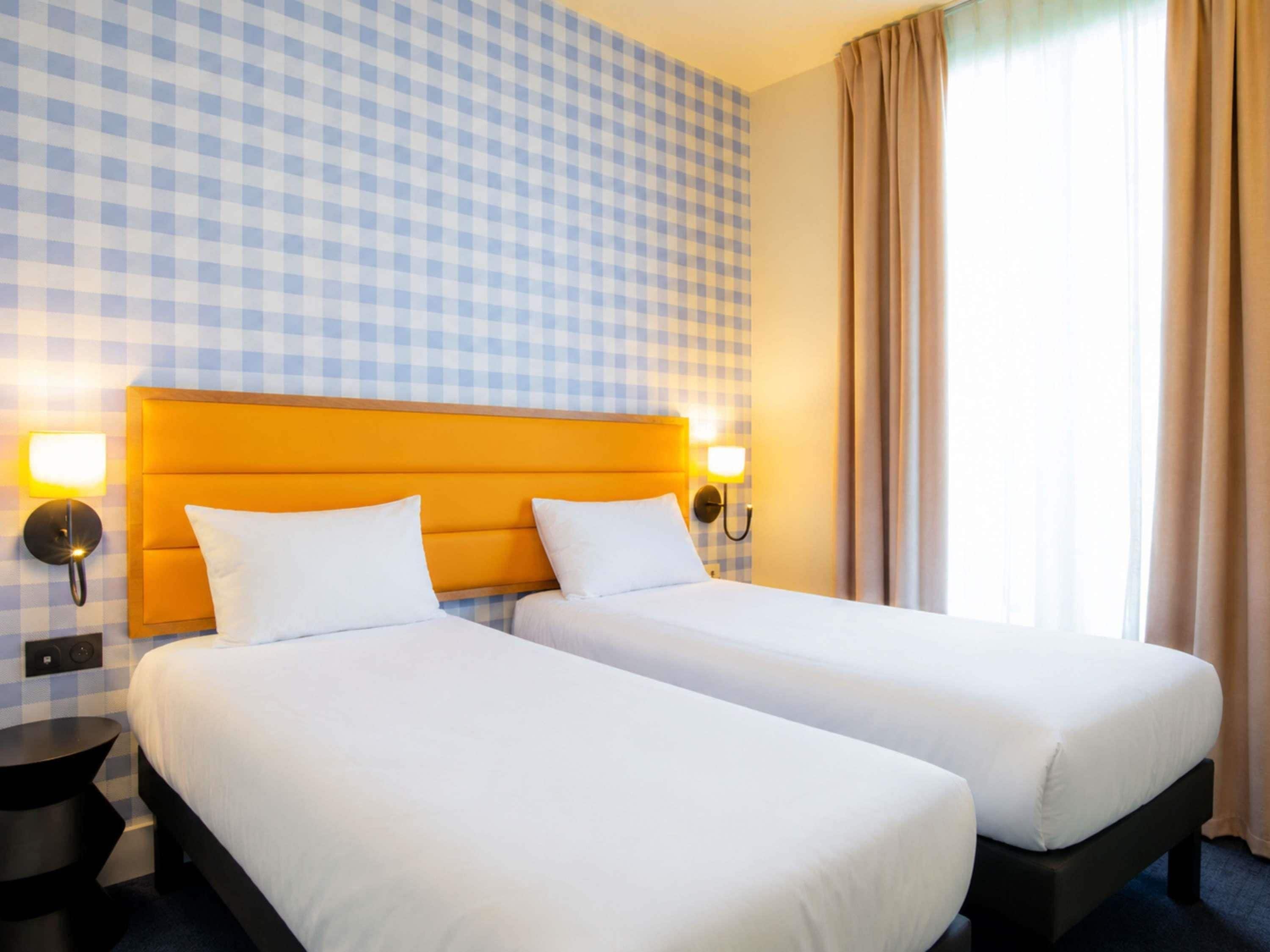 Une chambre d’hôtel avec deux lits, un papier peint à carreaux et une fenêtre avec des rideaux.