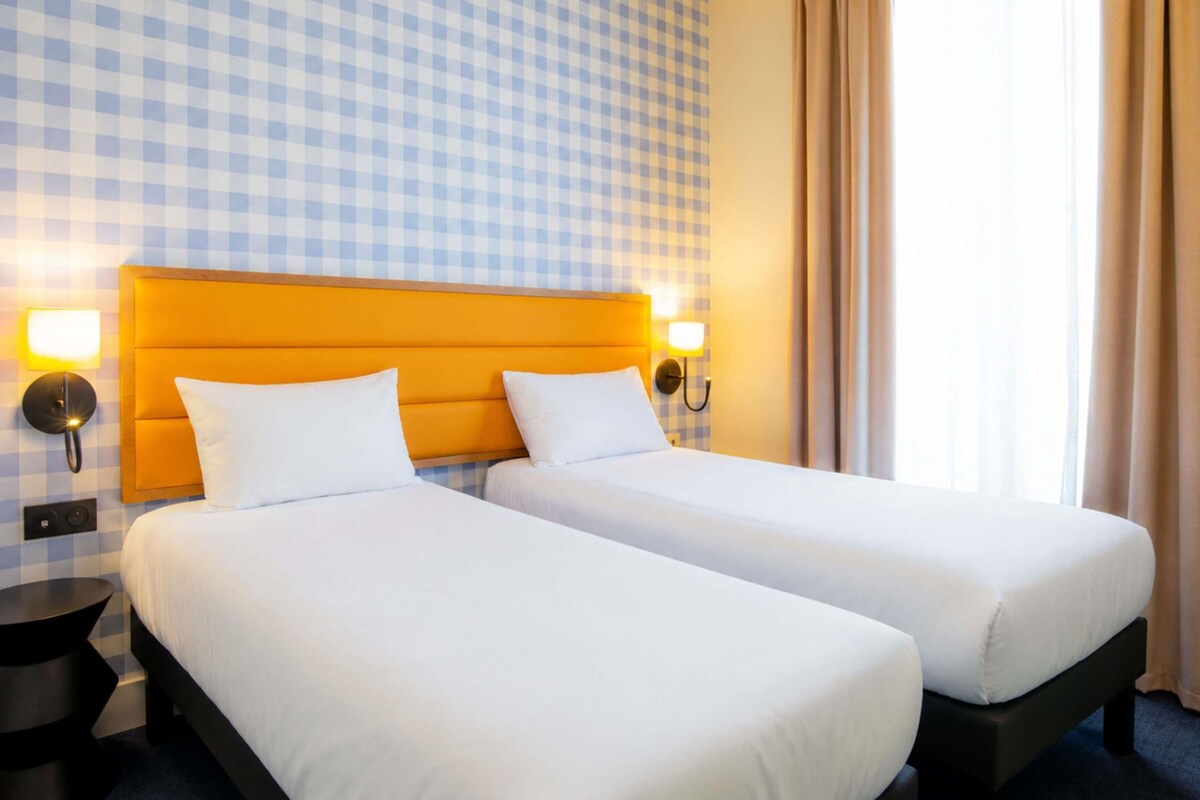 Una habitación de hotel con dos camas, papel tapiz a cuadros y un ventanal con cortinas.