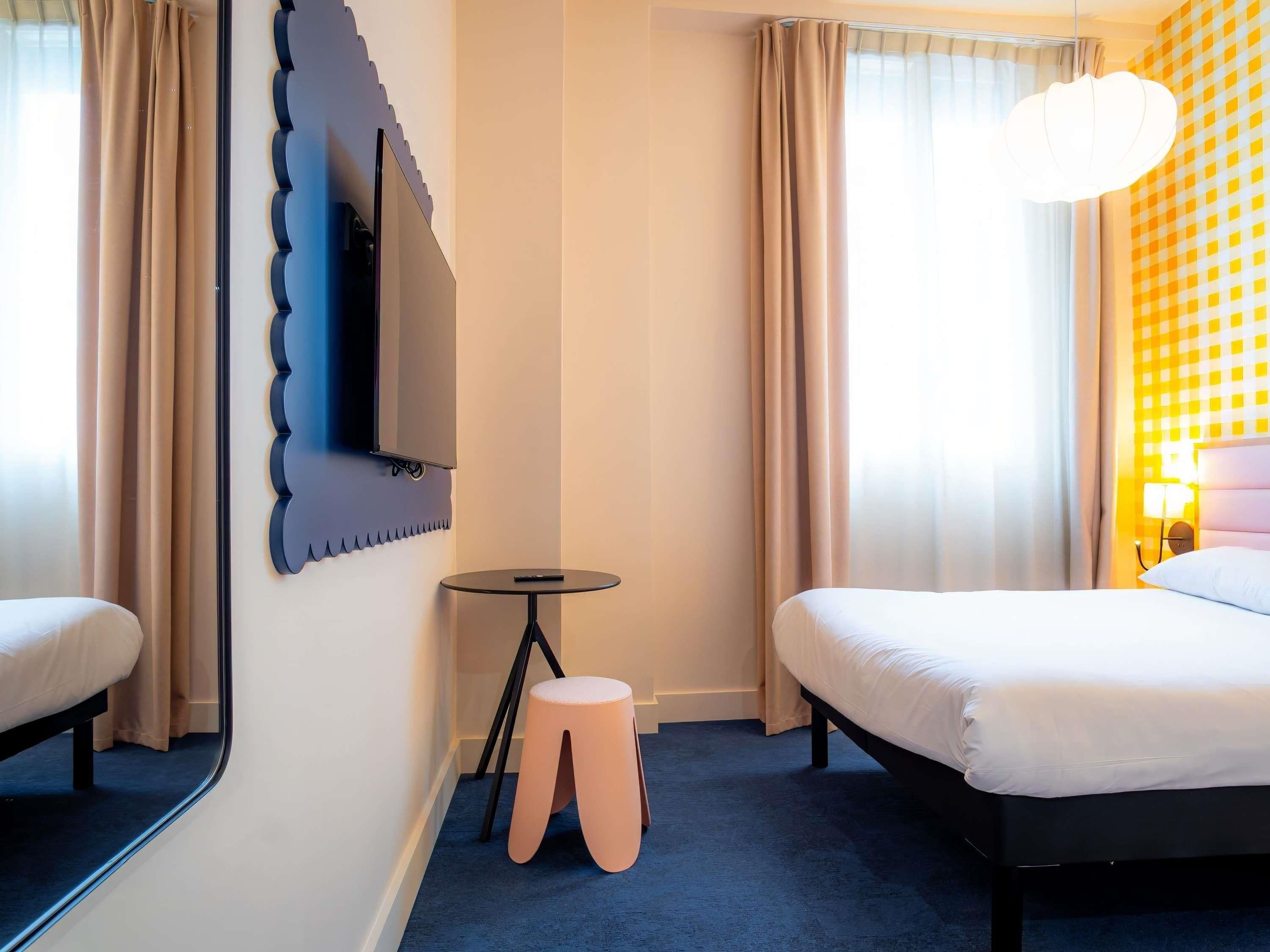 Habitación de hotel con dos camas, una mesita redonda pequeña, un taburete, un televisor montado en la pared y un papel tapiz estampado.