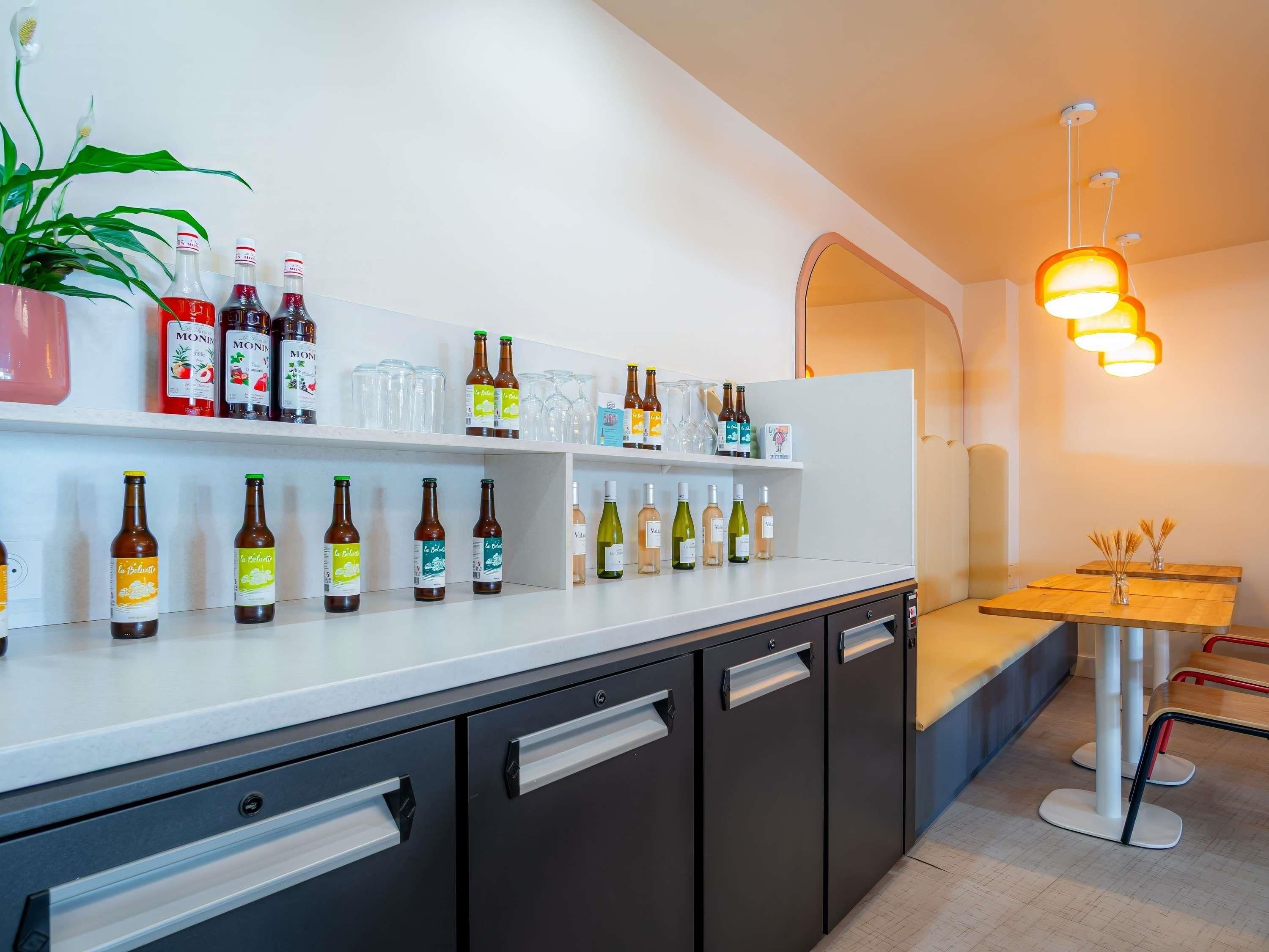 Zona de bar moderna con estantes llenos de bebidas embotelladas, una planta en maceta y un área de estar con mesa y sillas de madera.