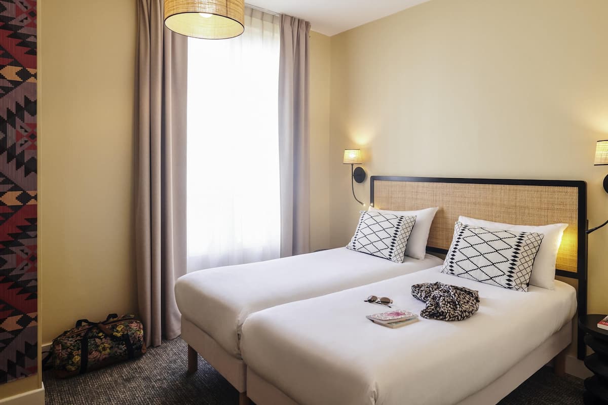 A hotel room with two beds, a hanging light fixture, a wall-mounted lamp, and a patterned curtain.