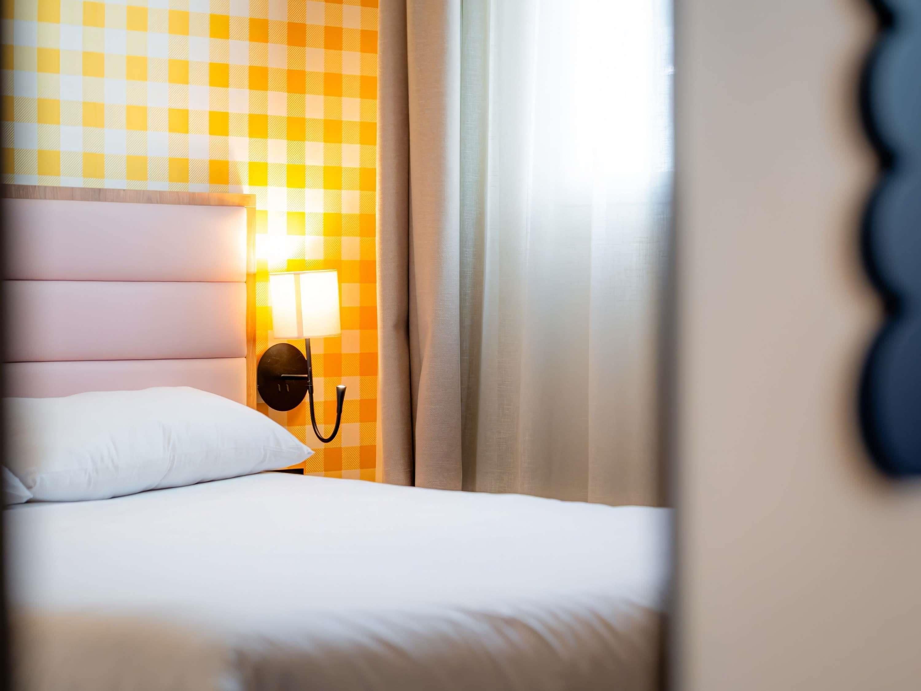 Quarto de hotel com parede xadrez, cama com roupa de cama branca e luminária fixada na parede.