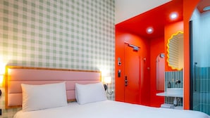 A hotel room with a checkered wall, a large bed with white bedding, a red accent wall, a modern bathroom with a mirror and sink, and a round bedside table.