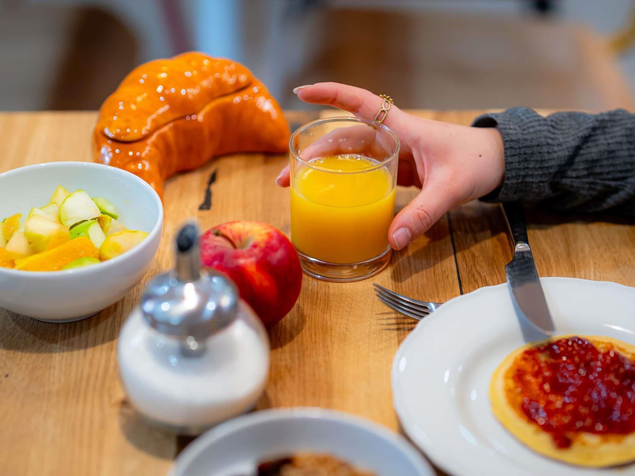 A breakfast setting with a bowl of fruit, a glass of orange juice, a croissant, a small jar, and a plate with jam.