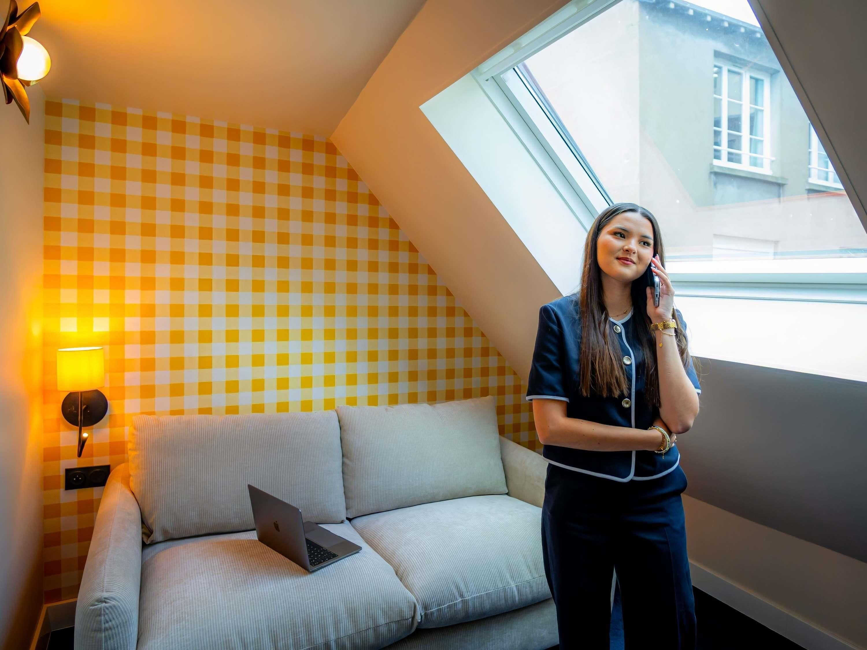 A room with a checkered wall, a sofa with a laptop, a lamp, and a window with a view of buildings.