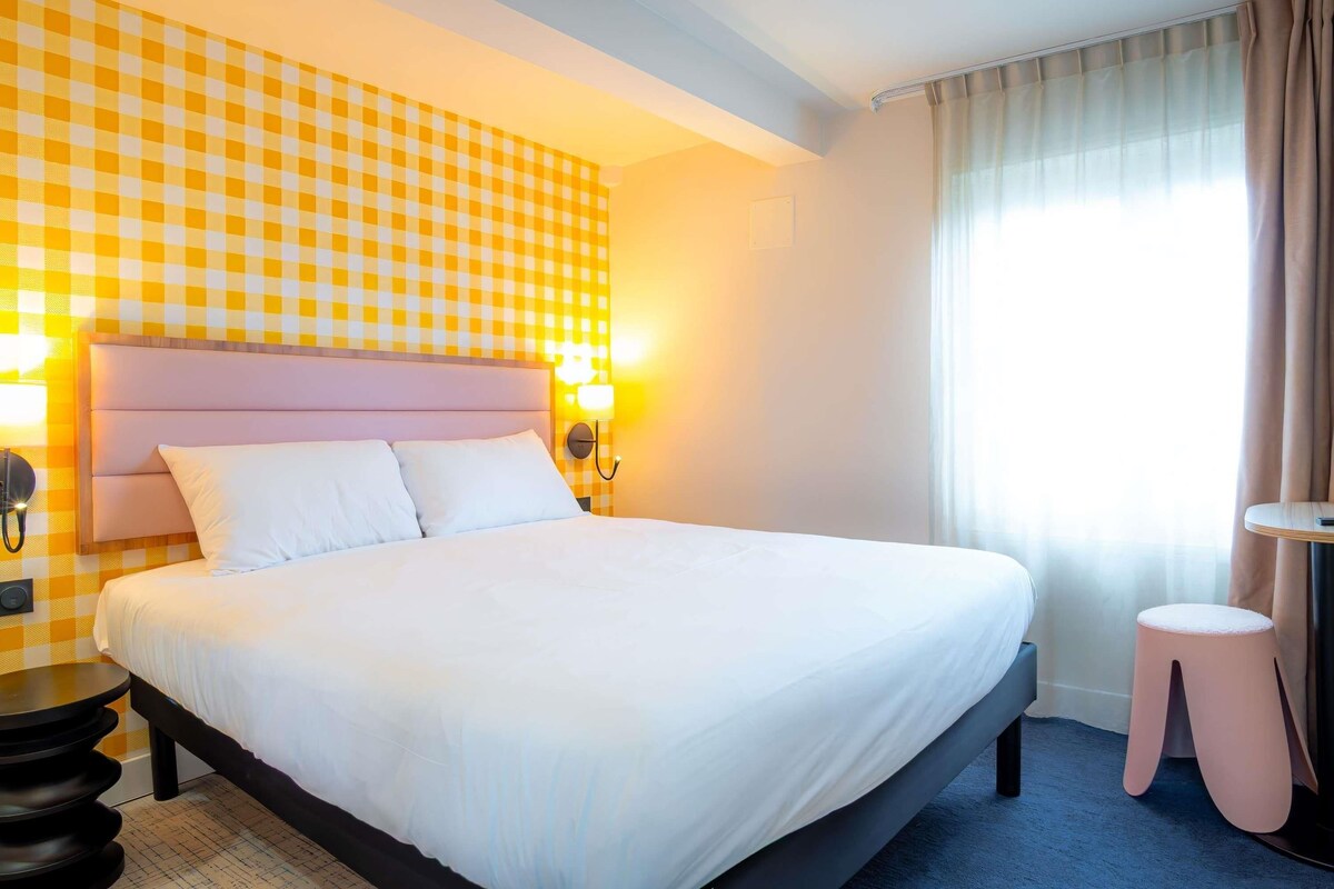 A hotel room with a checkered wall, a bed with white linens, a headboard, bedside lamps, a small round table, and a stool.