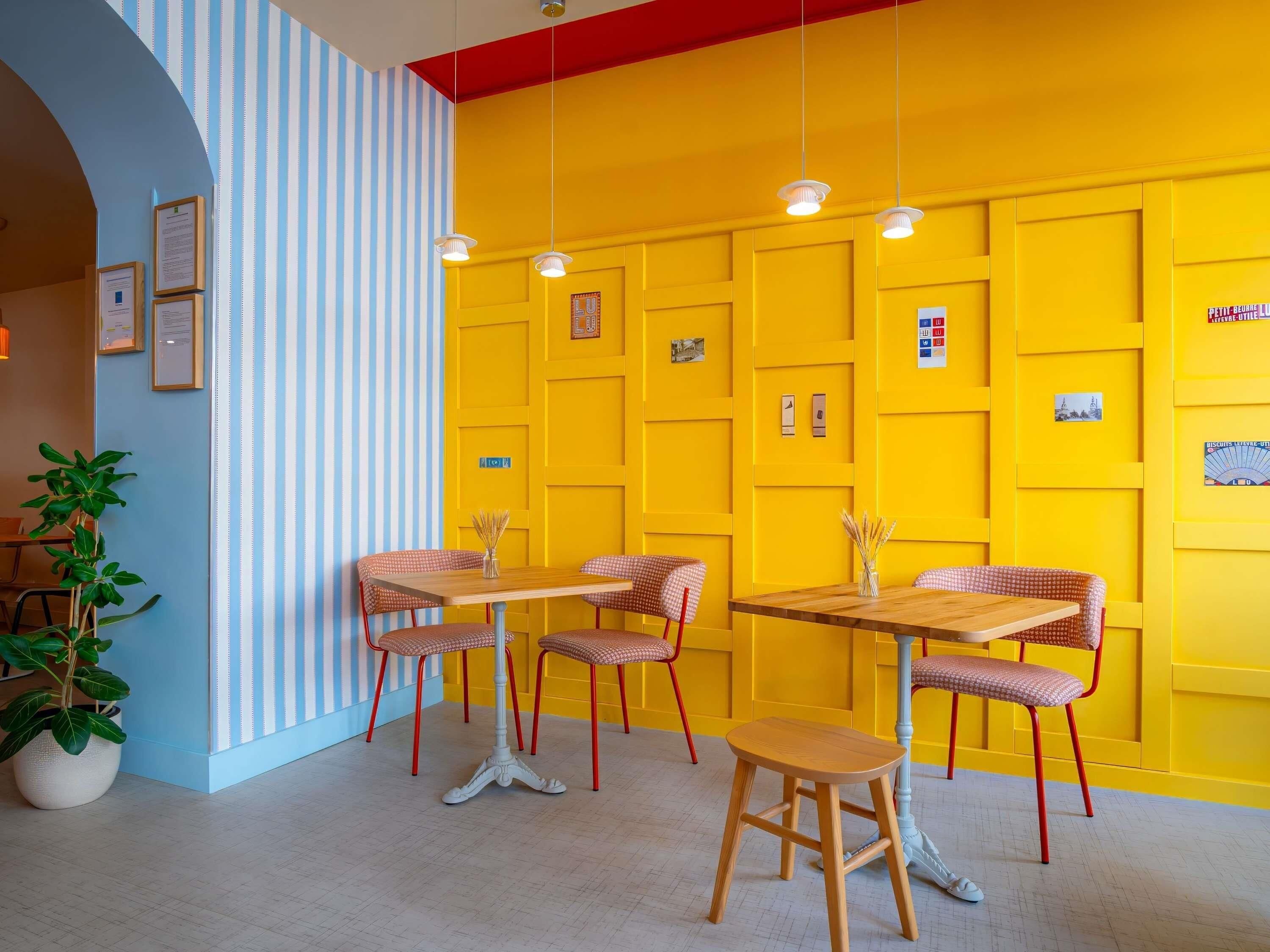 A room with a yellow wall, striped blue and white wall, and a white ceiling. There are wooden tables and chairs, a potted plant, and hanging lights.