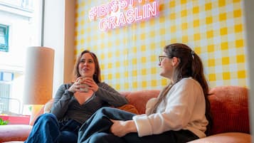Two people sitting on a couch with a checkered wall and a neon sign in the background.