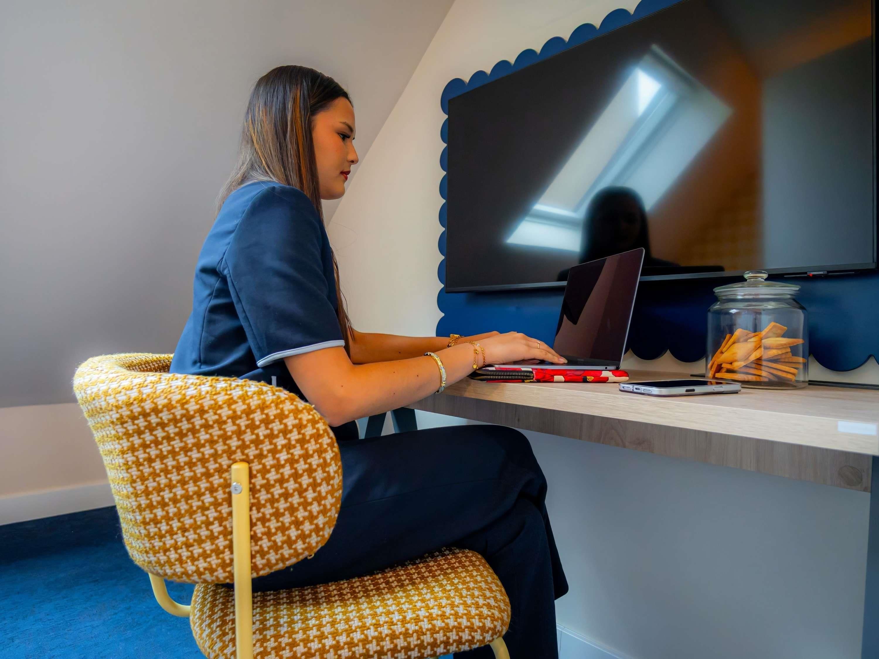 A person seated on a patterned chair at a desk with a laptop, a smartphone, and a jar of snacks.