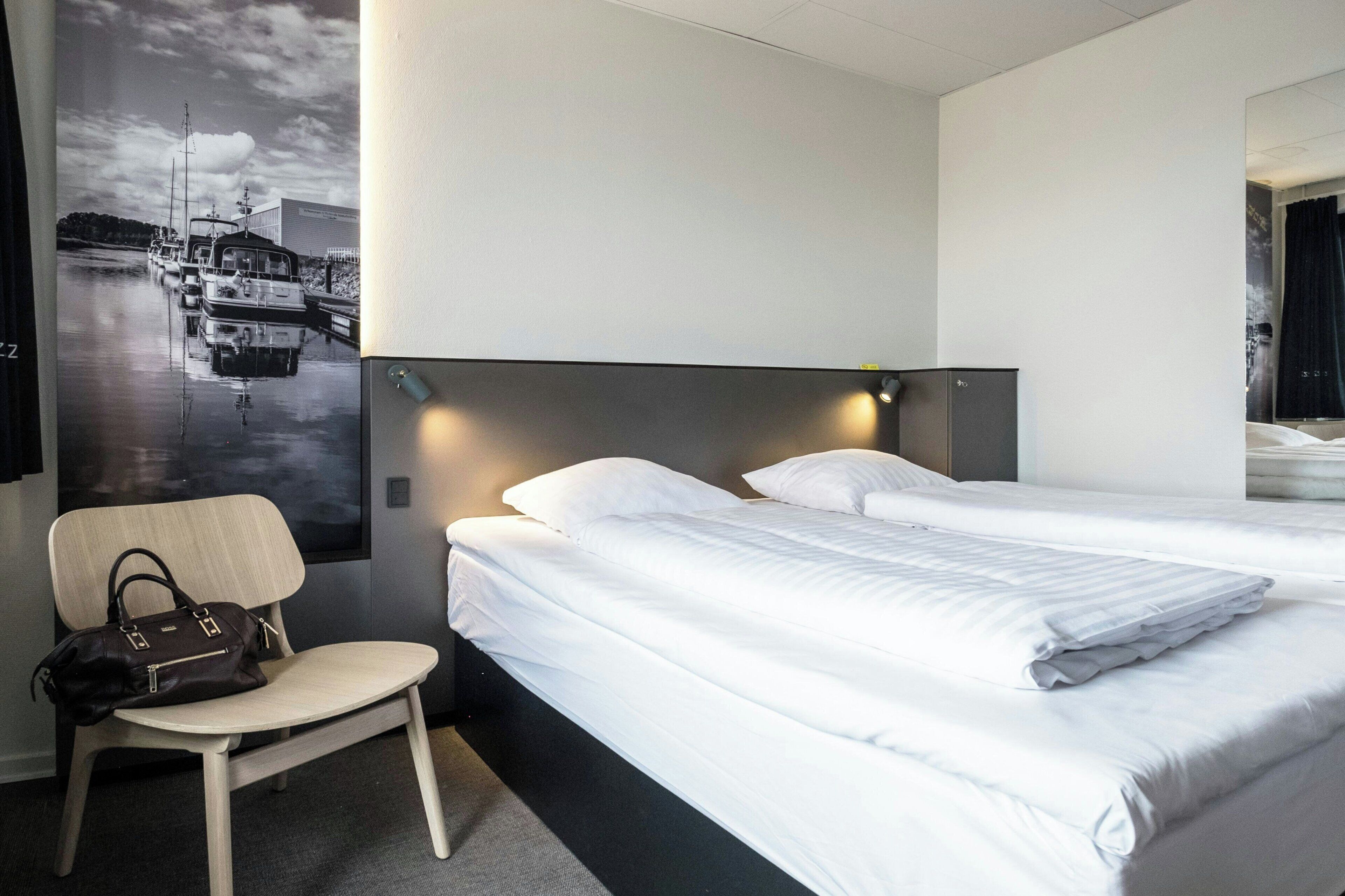 Quarto de hotel com cama, uma cadeira e cabeceira fixada na parede.