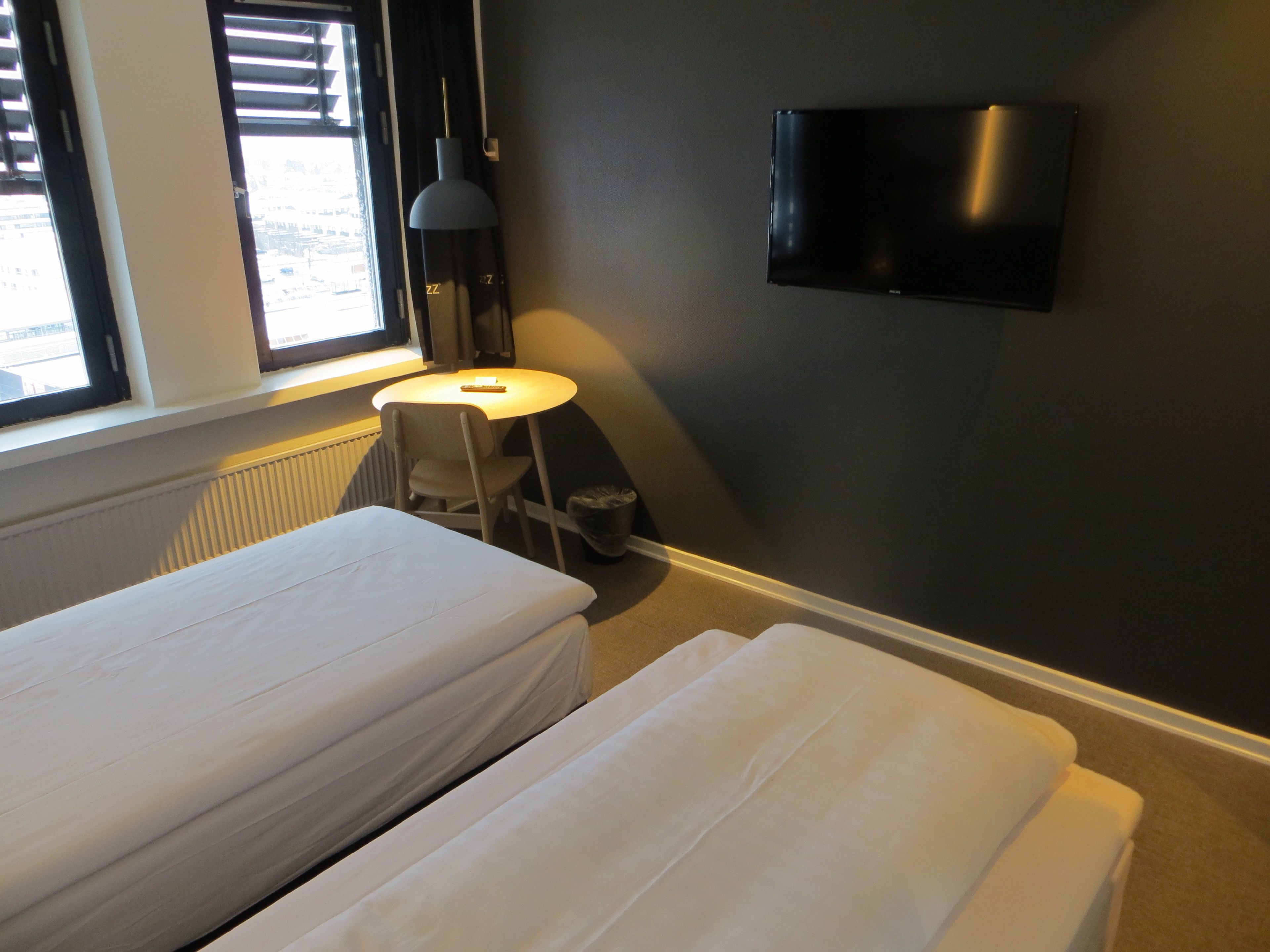 Habitación de hotel con dos camas individuales, una mesita redonda pequeña, una silla, un televisor montado en la pared y una ventana con persianas.