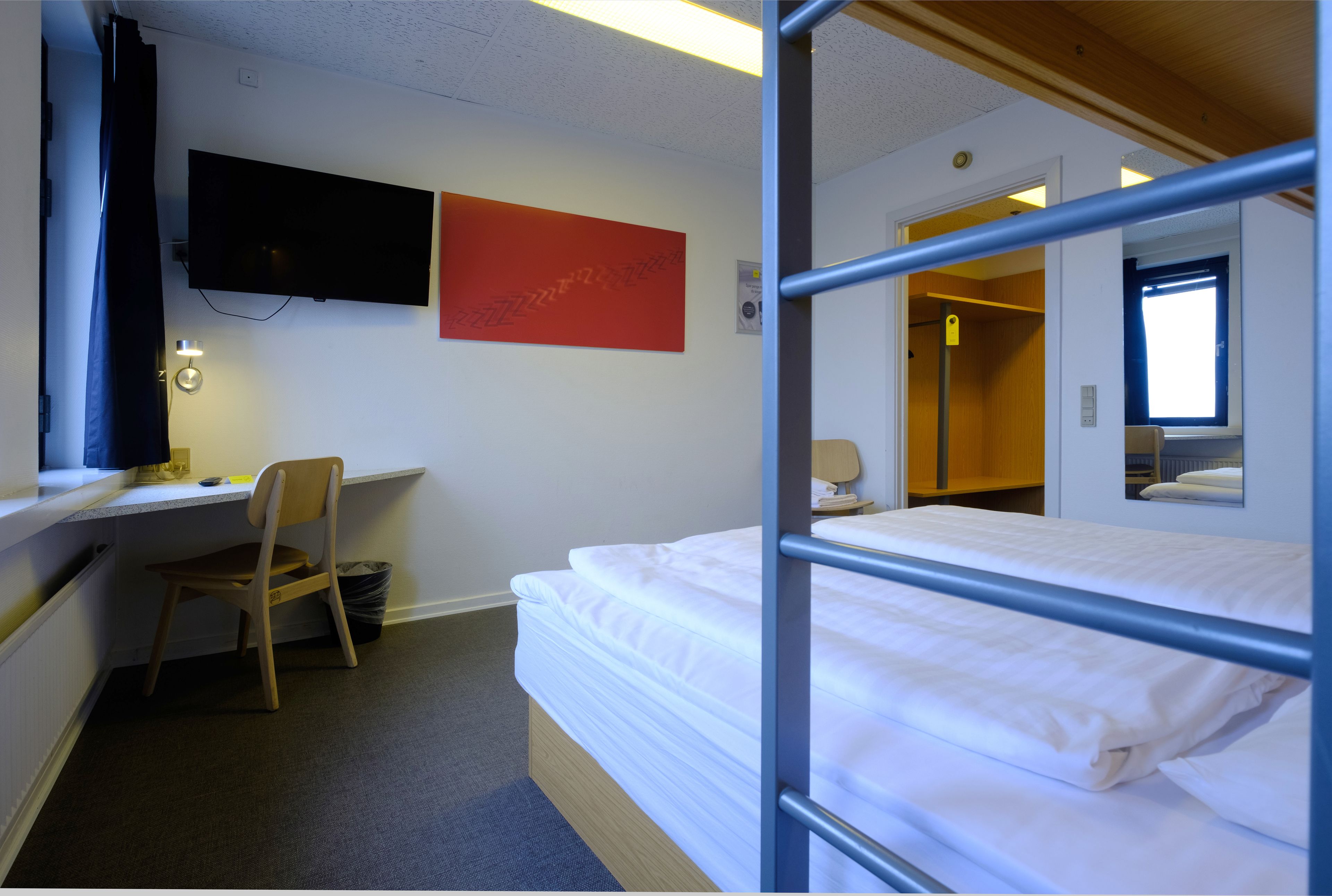A room with a bunk bed, a desk with a chair, a television mounted on the wall, and a red abstract painting.