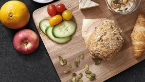 Tagliere di legno con cetrioli a fette, pomodorini, una mela, una fetta di formaggio, un panino e semi di zucca.
