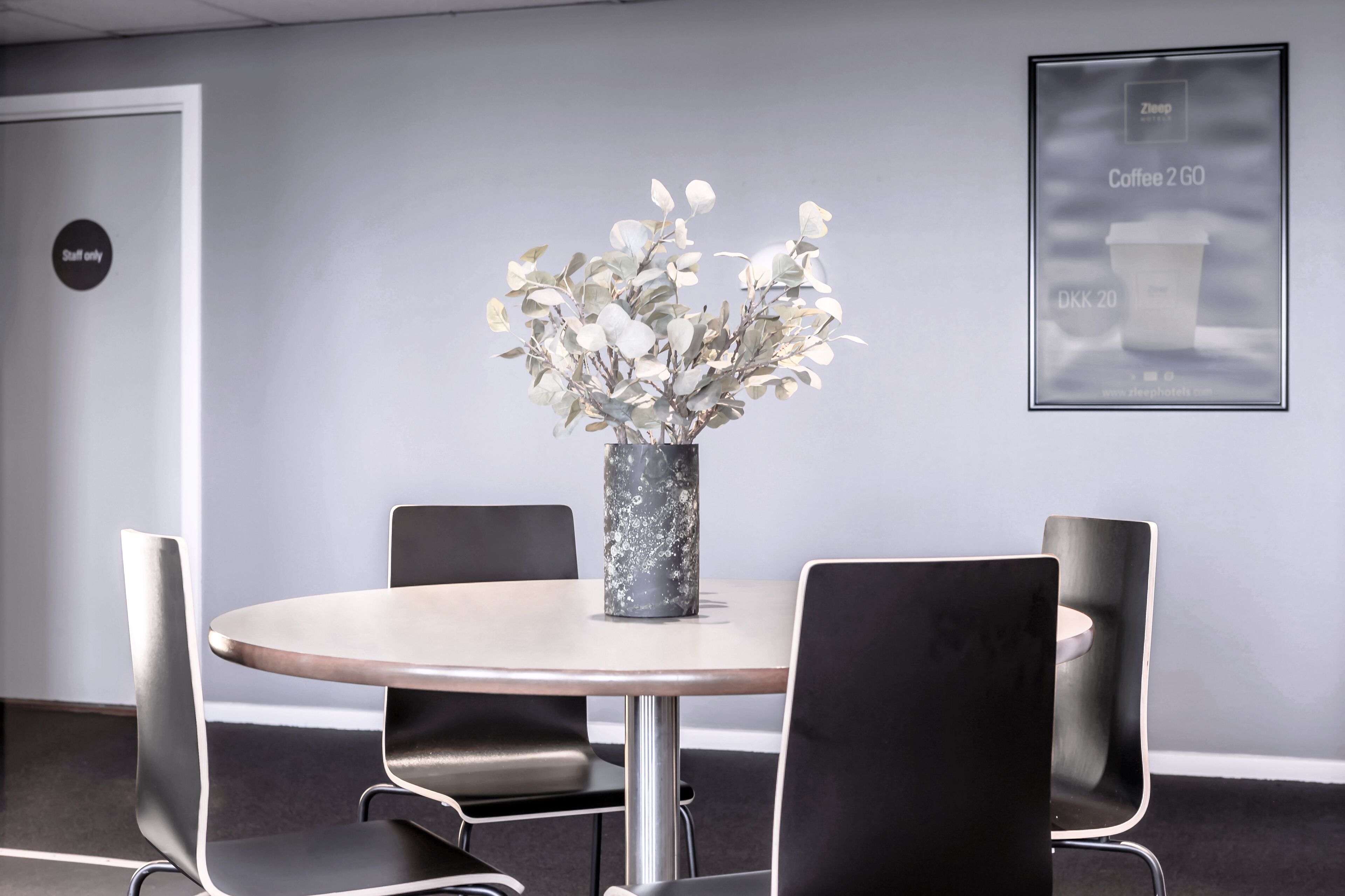 Un salón de conferencias con una mesa redonda, cuatro sillas negras, un jarrón con flores blancas y un cartel enmarcado en la pared.