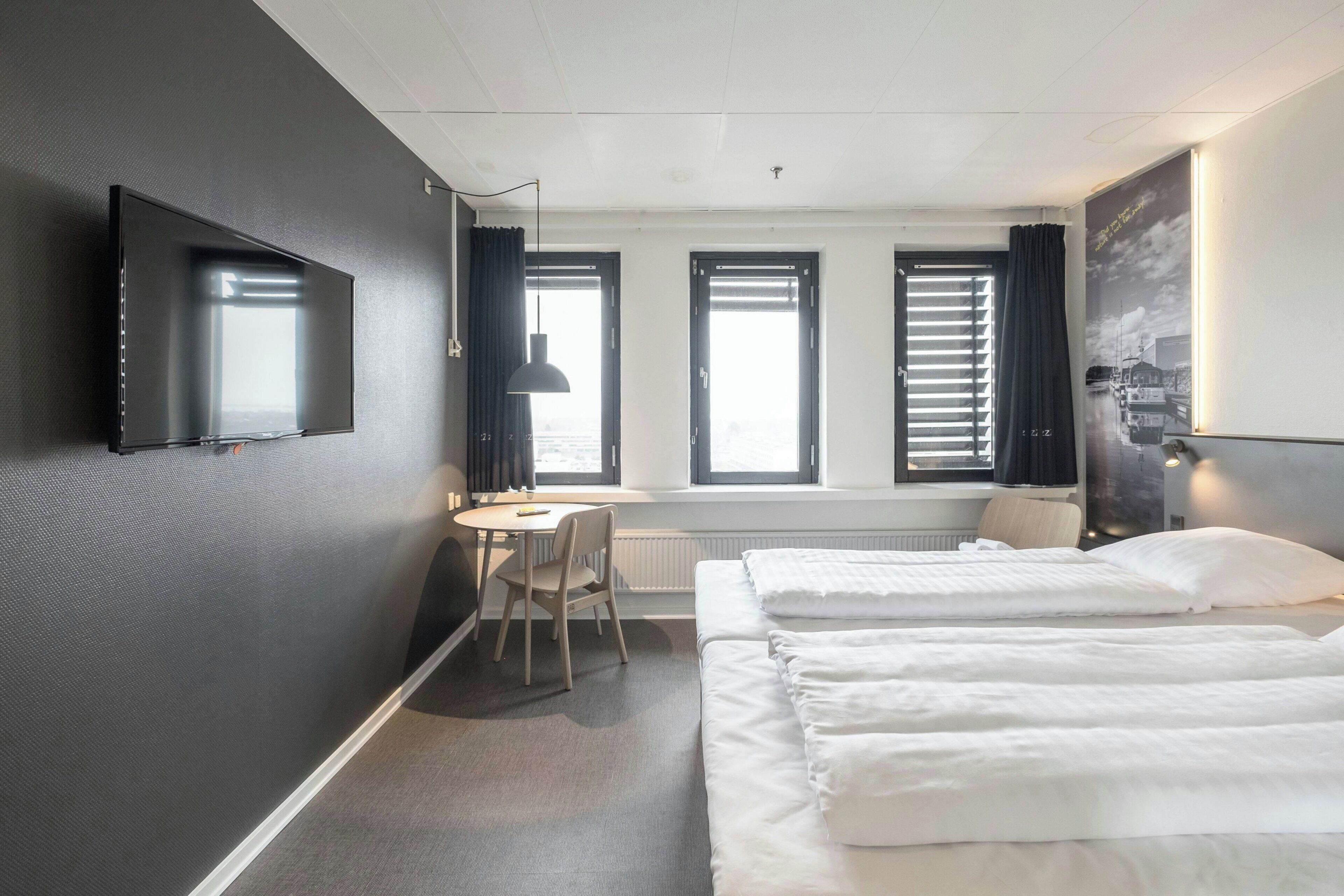 Hotellihuone, jossa on seinälle asennettu taulutelevisio, työpöytä tuolilla, kaksi sänkyä ja suuret ikkunat, joissa on sälekaihtimet.