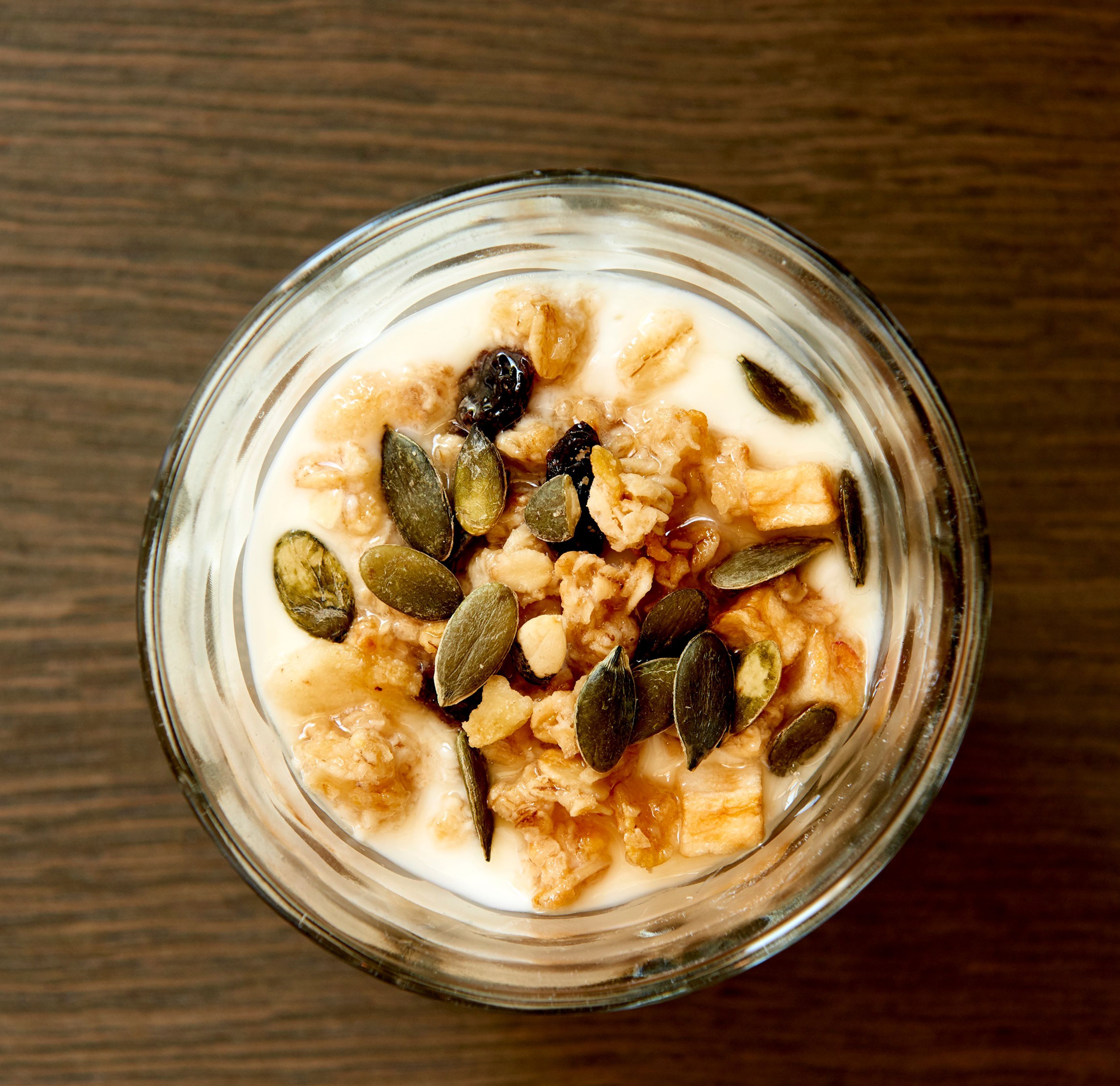 A glass bowl of cereal with milk and pumpkin seeds on a wooden surface.