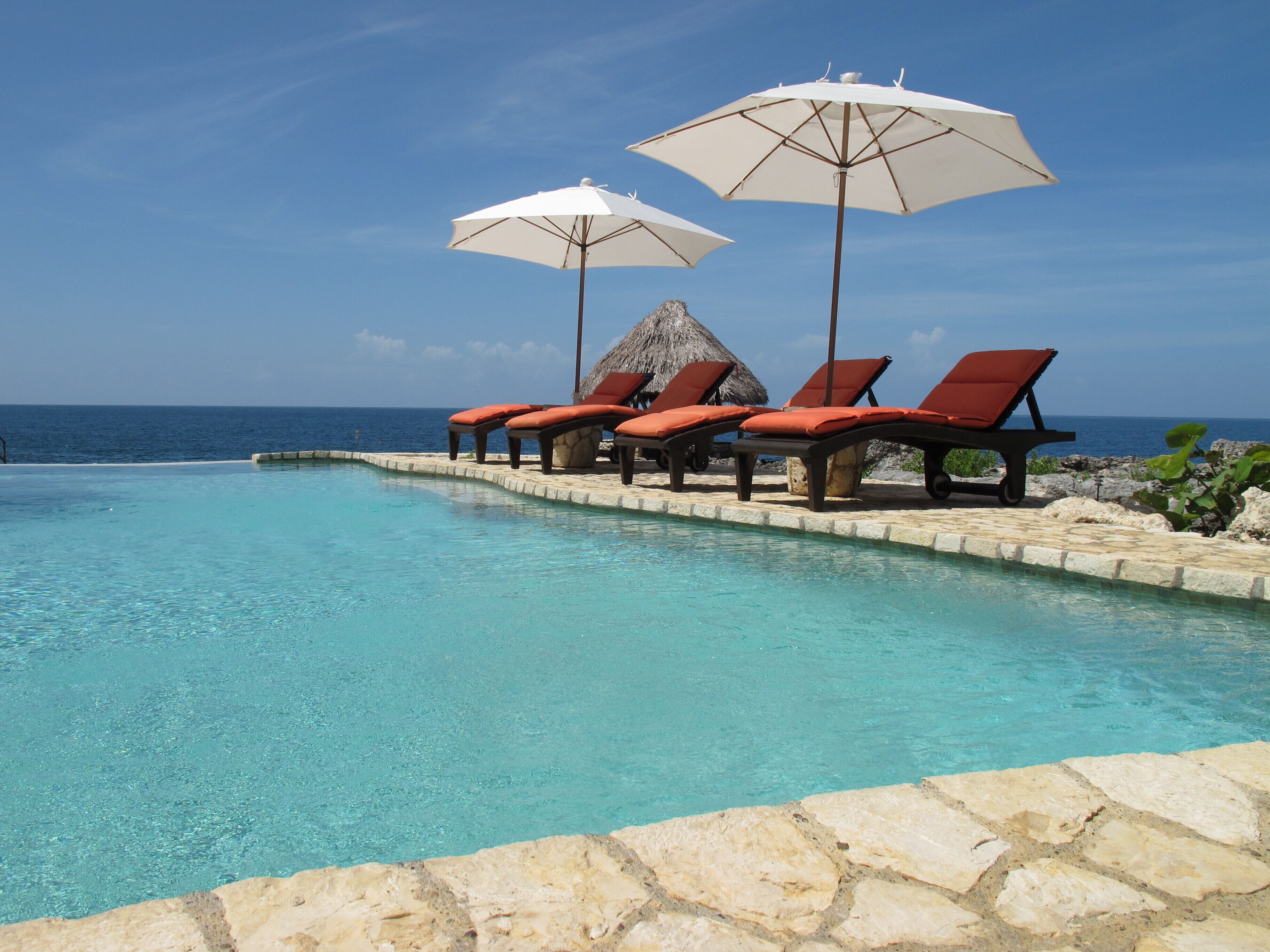 A poolside area with lounge chairs and umbrellas, a stone border, and a view of the ocean.