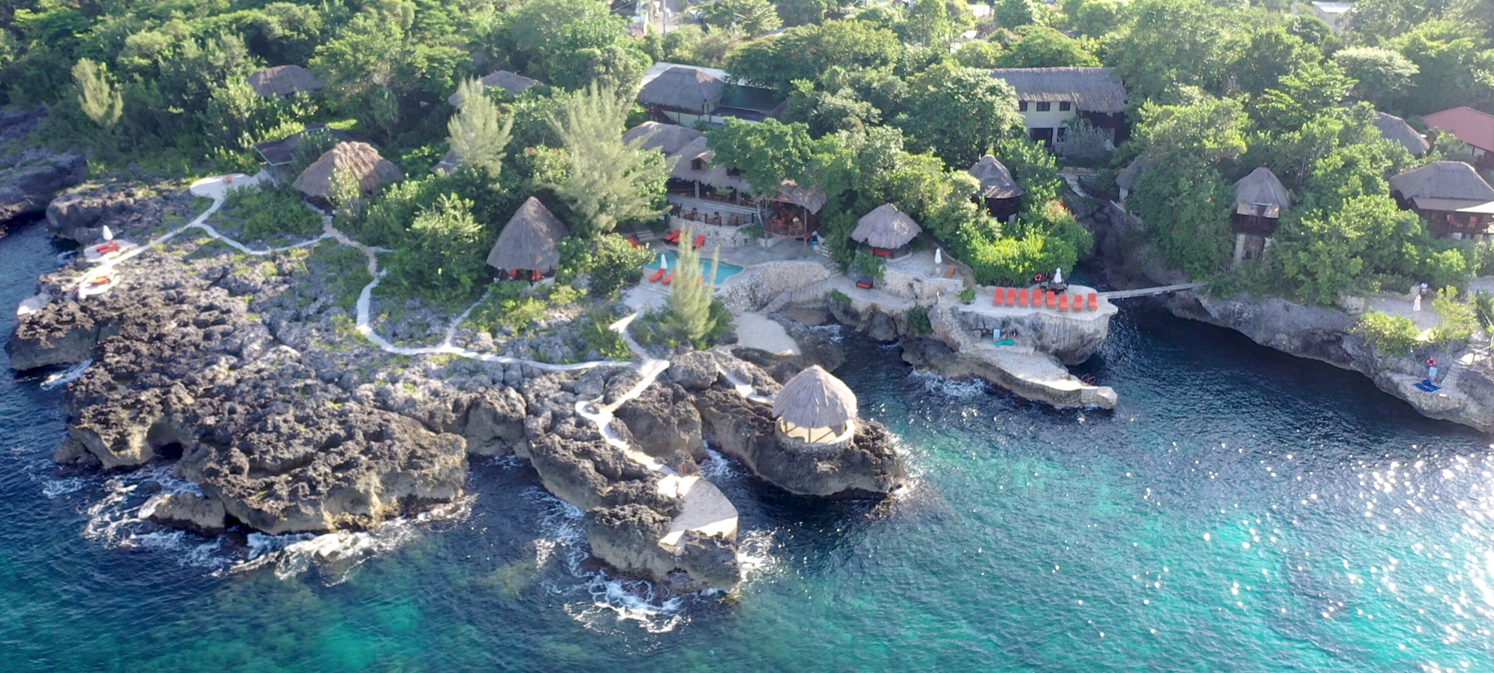 Vista aérea de un resort costero con cabañas de techo de paja, aguas turquesas cristalinas y un litoral rocoso.