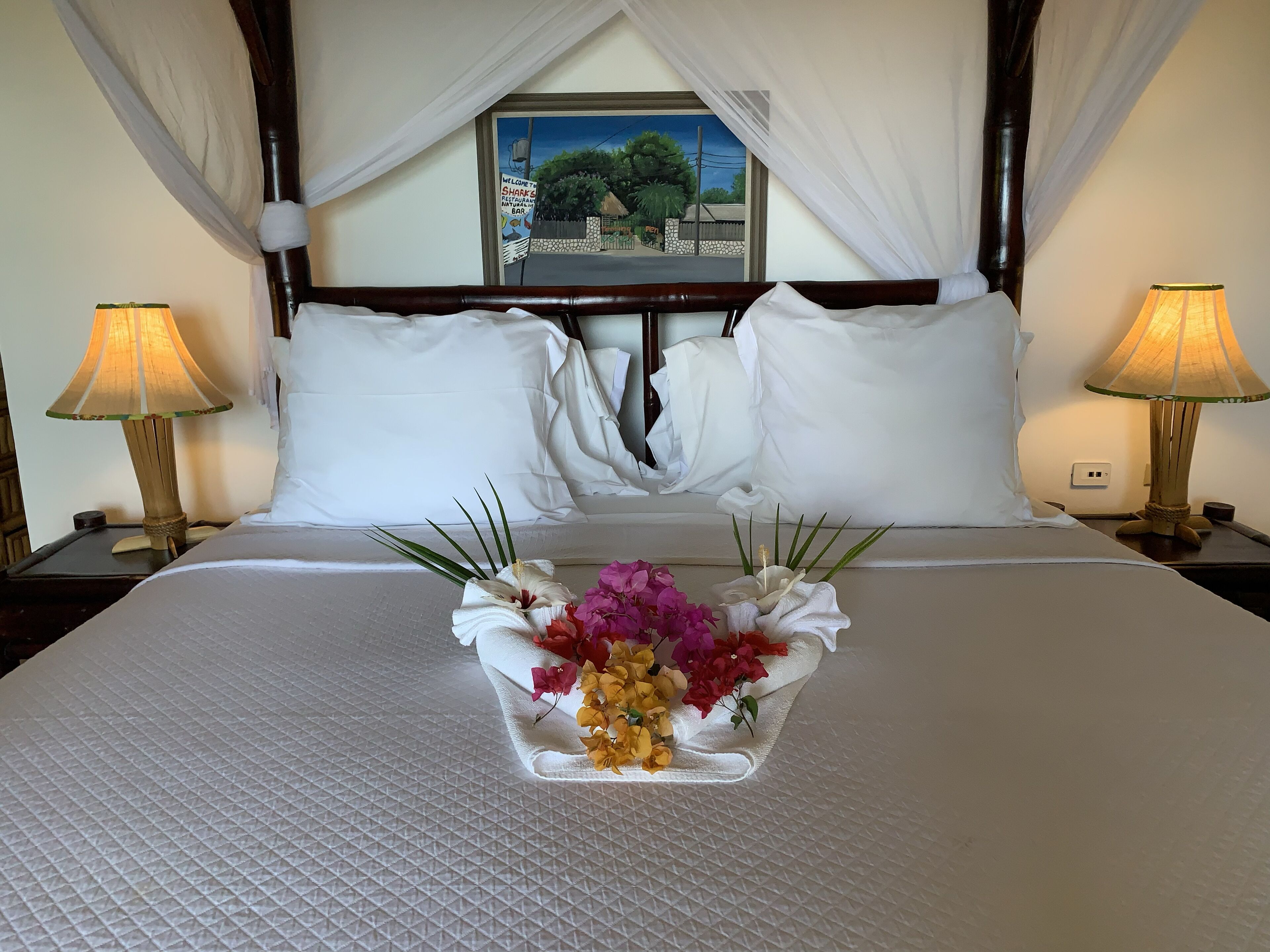 Un lit bien fait, recouvert d’une courtepointe blanche, avec deux oreillers blancs et un arrangement floral posé sur le lit. Deux lampes de chevet sont placées de chaque côté du lit.