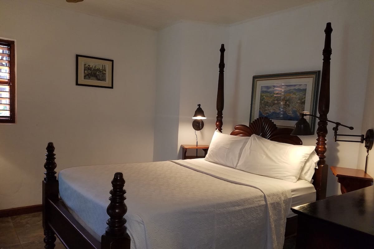 Una cama con dosel con cubrecama blanco, una mesita de noche de madera con lámpara y un cuadro enmarcado en la pared.