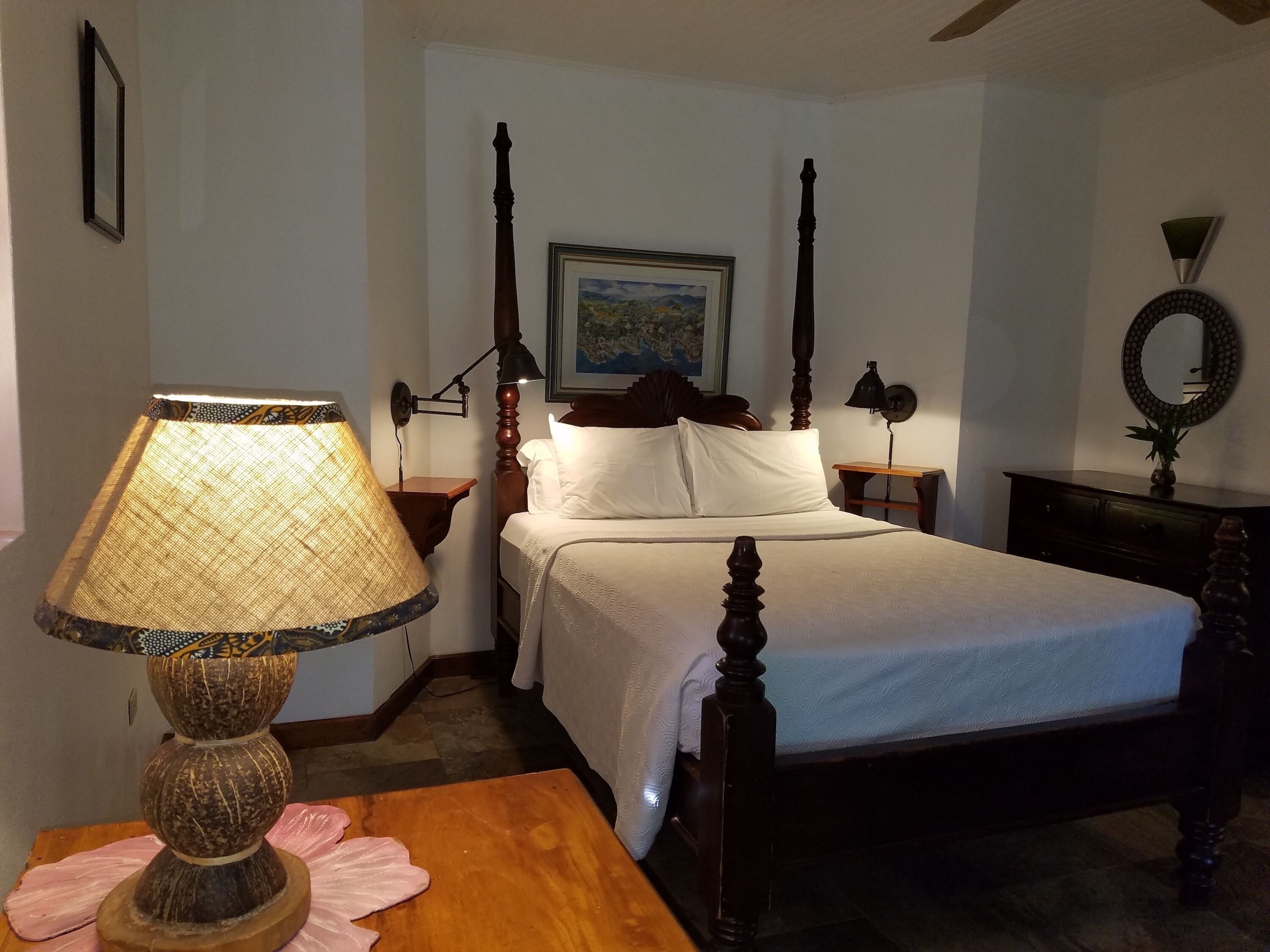 A bedroom with a four-poster bed, a bedside lamp, a framed painting on the wall, and a wooden dresser.