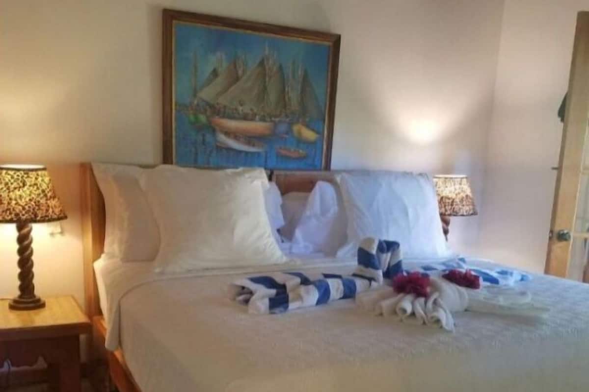 Una cama bien hecha con sábanas blancas y una manta a rayas, dos lámparas de noche y un cuadro de un velero en la pared.
