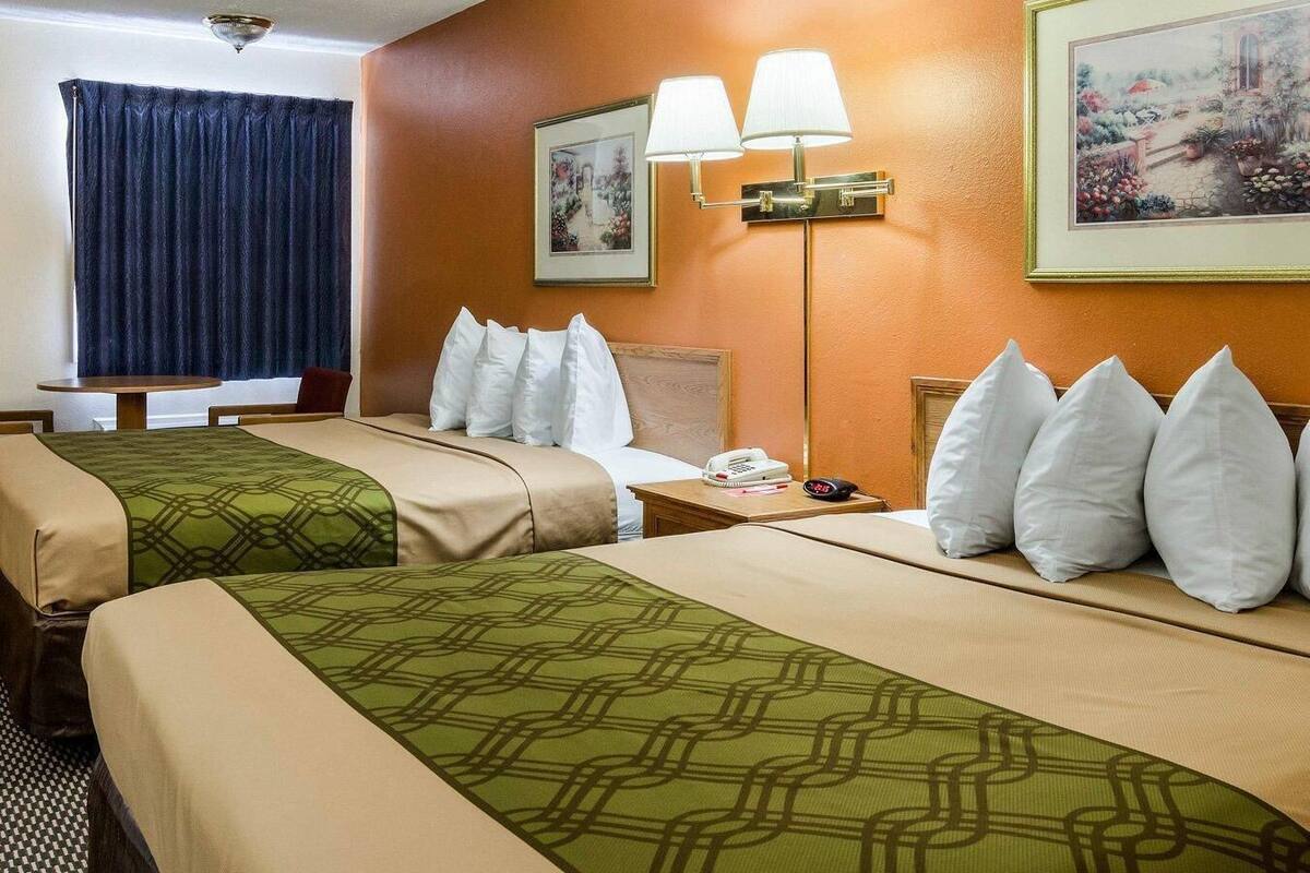 A hotel room with two beds, a bedside table with a lamp, a painting on the wall, and a window with curtains.