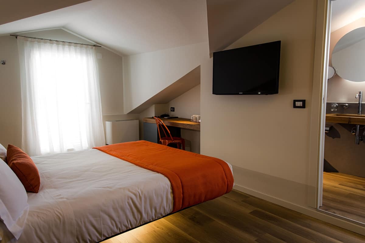 A bedroom with a bed, a desk, a chair, a TV, and a bathroom visible through a glass door.