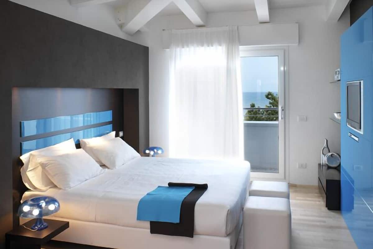 A modern bedroom with a large bed, a bedside table with a lamp, a small bench, and a view of the outdoors.
