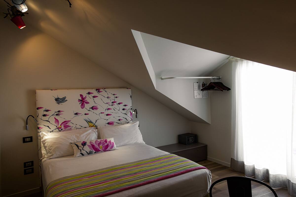 A bedroom with a bed, a nightstand, a wall-mounted shelf, and a window with curtains.