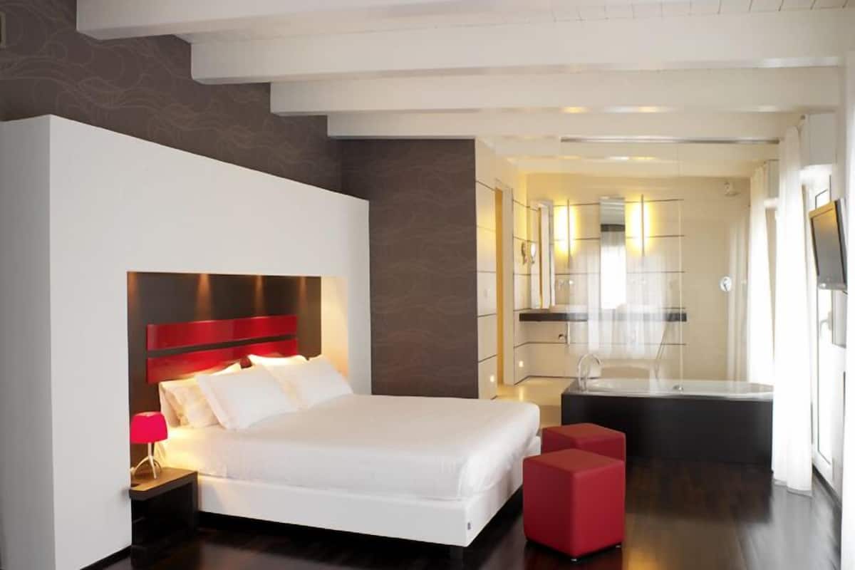 A modern hotel room with a large bed, a red accent wall, a separate seating area, and a bathroom visible through an open door.