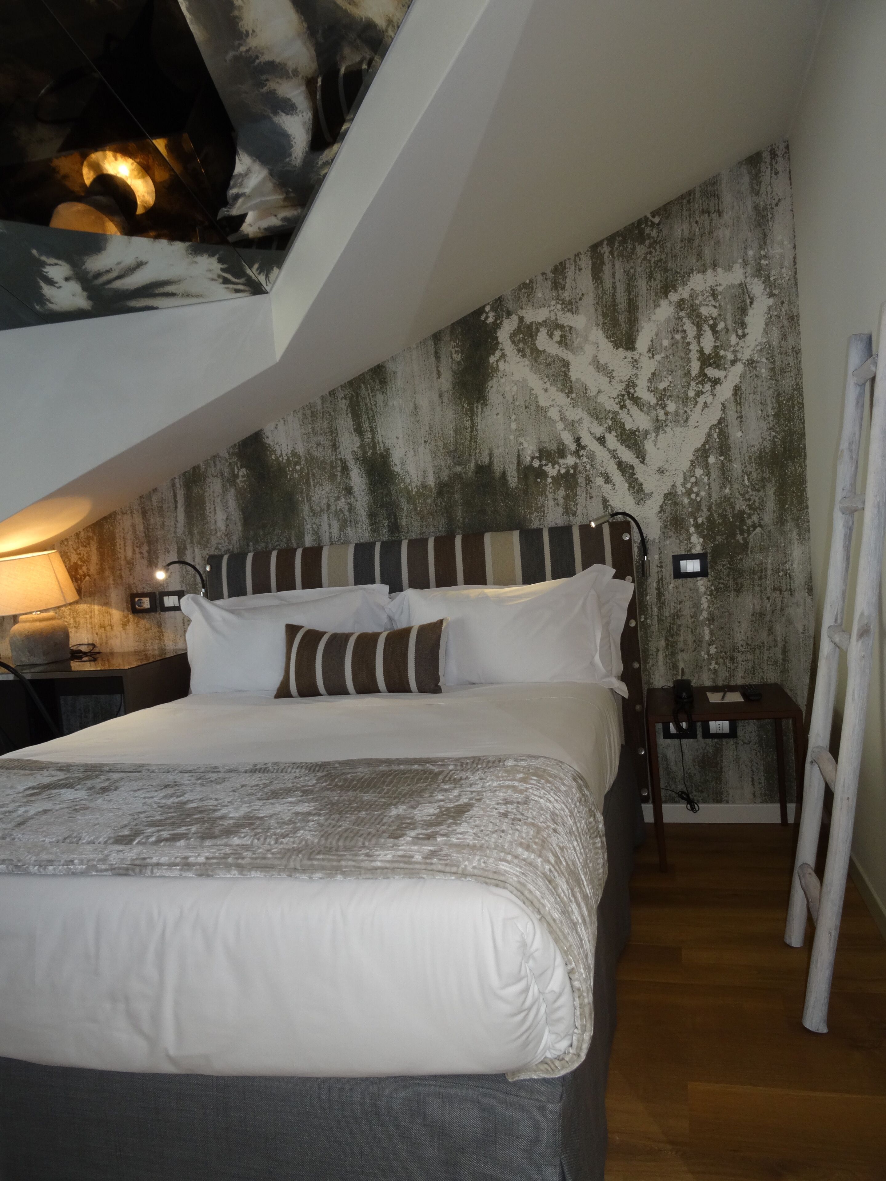 A bedroom with a sloped ceiling, a bed with white linens, a striped pillow, a nightstand with a lamp, and a wall with a textured pattern.