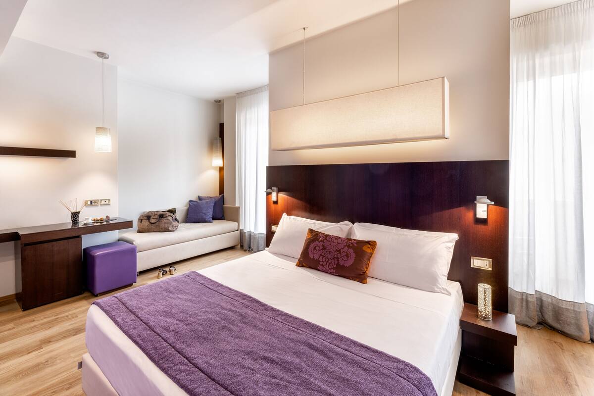 A modern hotel room with a large bed, a desk, a sofa, and a nightstand with a lamp.