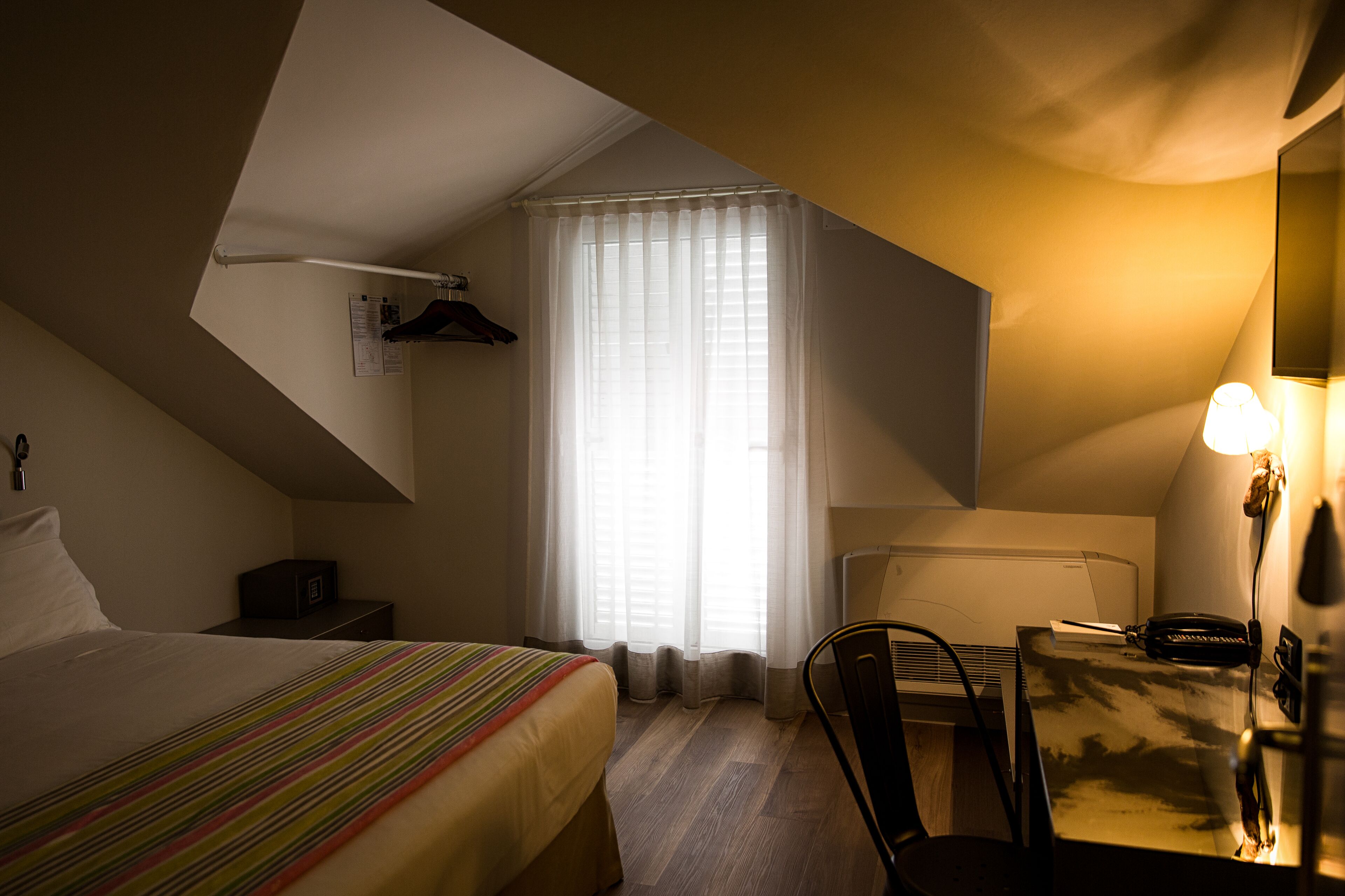 Una habitación de hotel con cama, escritorio, silla, lámpara y ventana con cortinas.