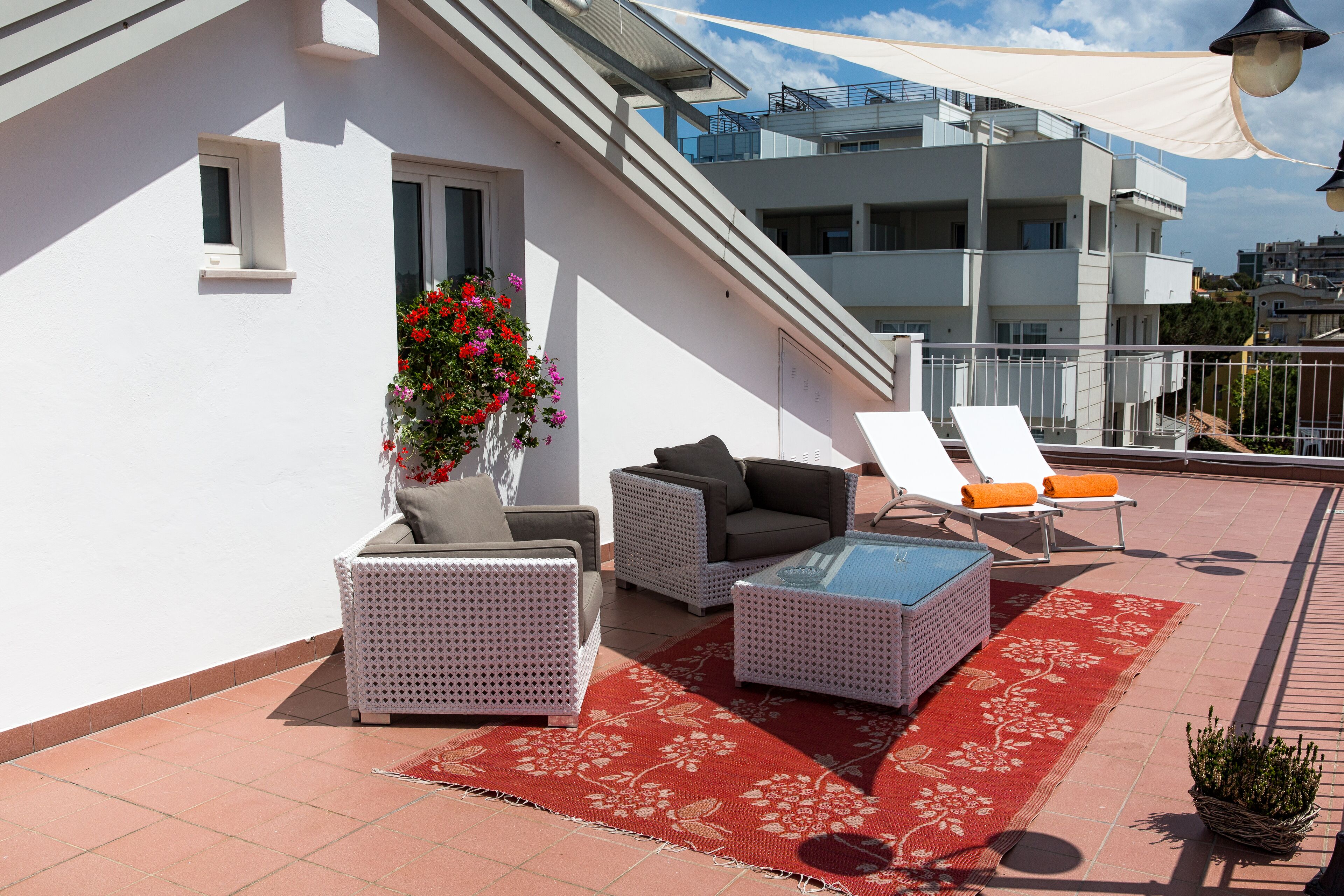 Eine Dachterrasse mit weißen Korbmöbeln, einem roten floralen Teppich und einem Blumentopf.