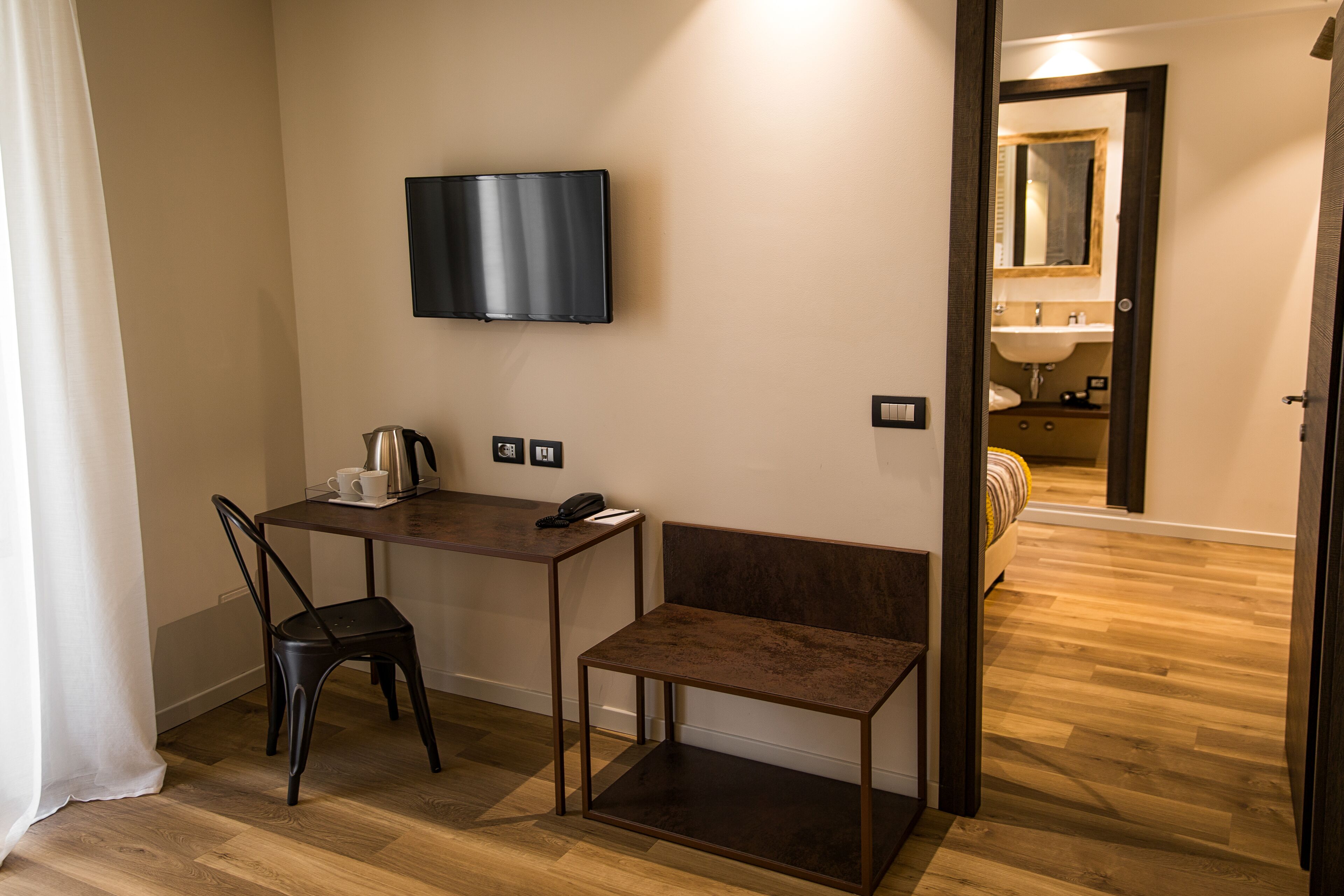 Habitación de hotel con piso de madera, un escritorio con silla, un televisor montado en la pared, un pequeño banco y una puerta que da a un baño.