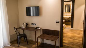 Habitación de hotel con piso de madera, un escritorio con silla, un televisor montado en la pared, un pequeño banco y una puerta que da a un baño.