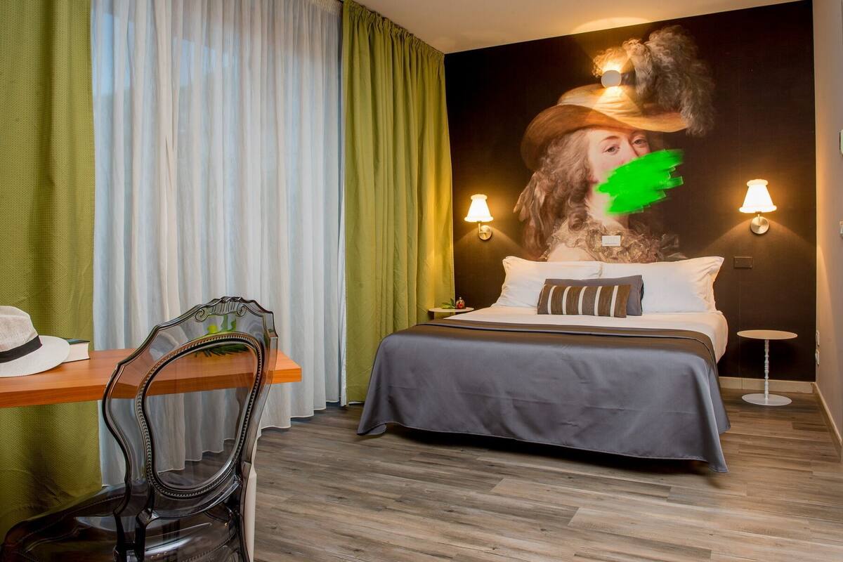 A hotel room with a bed, a chair, a table, and a wall with a painting.
