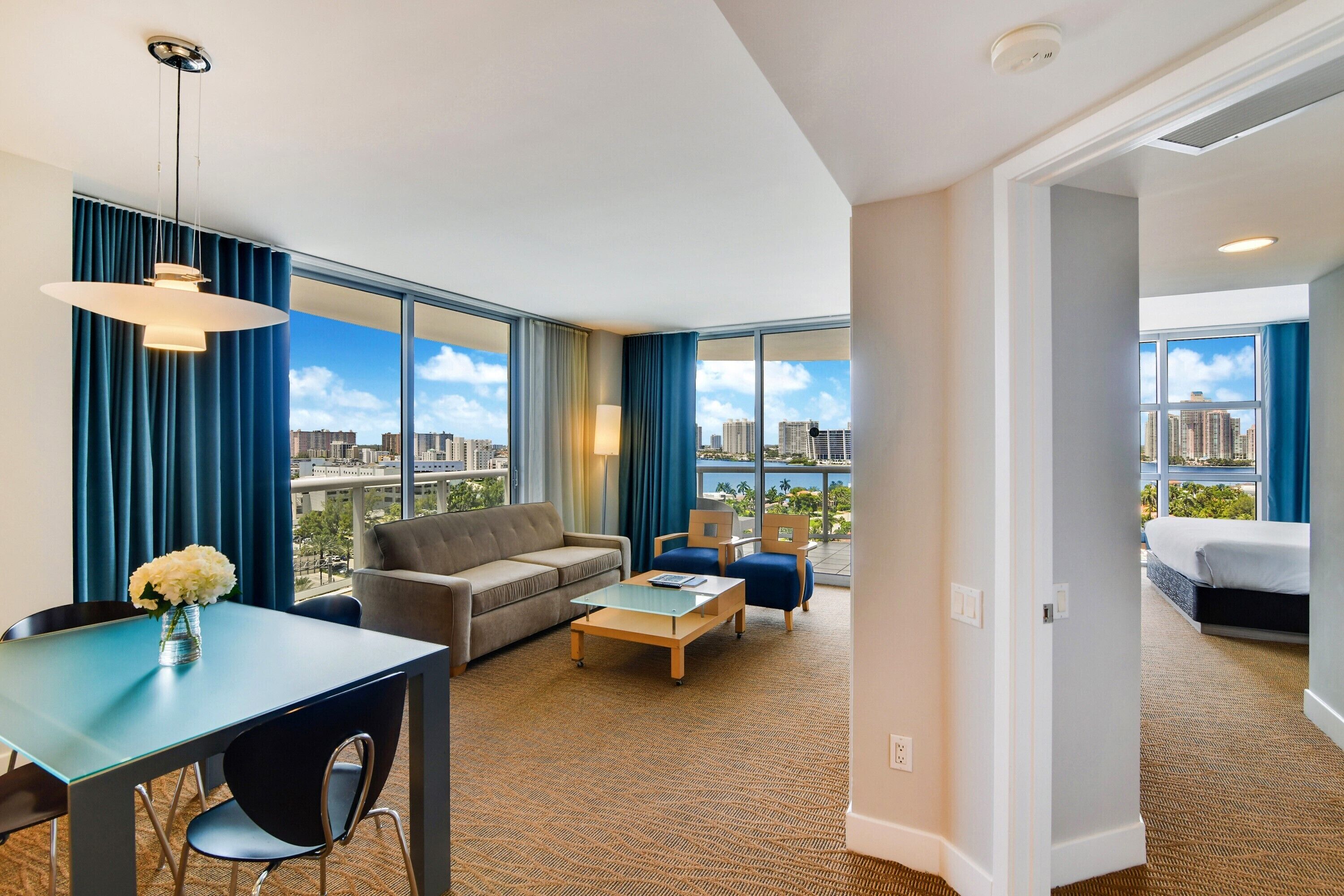 A modern hotel room with a large window, a sofa, a coffee table, and a dining area.