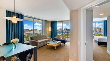 A modern hotel room with a large window, a sofa, a coffee table, and a dining area.