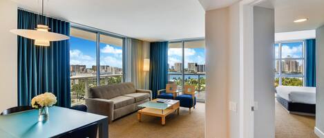 A modern hotel room with a large window, a sofa, a coffee table, and a dining area.