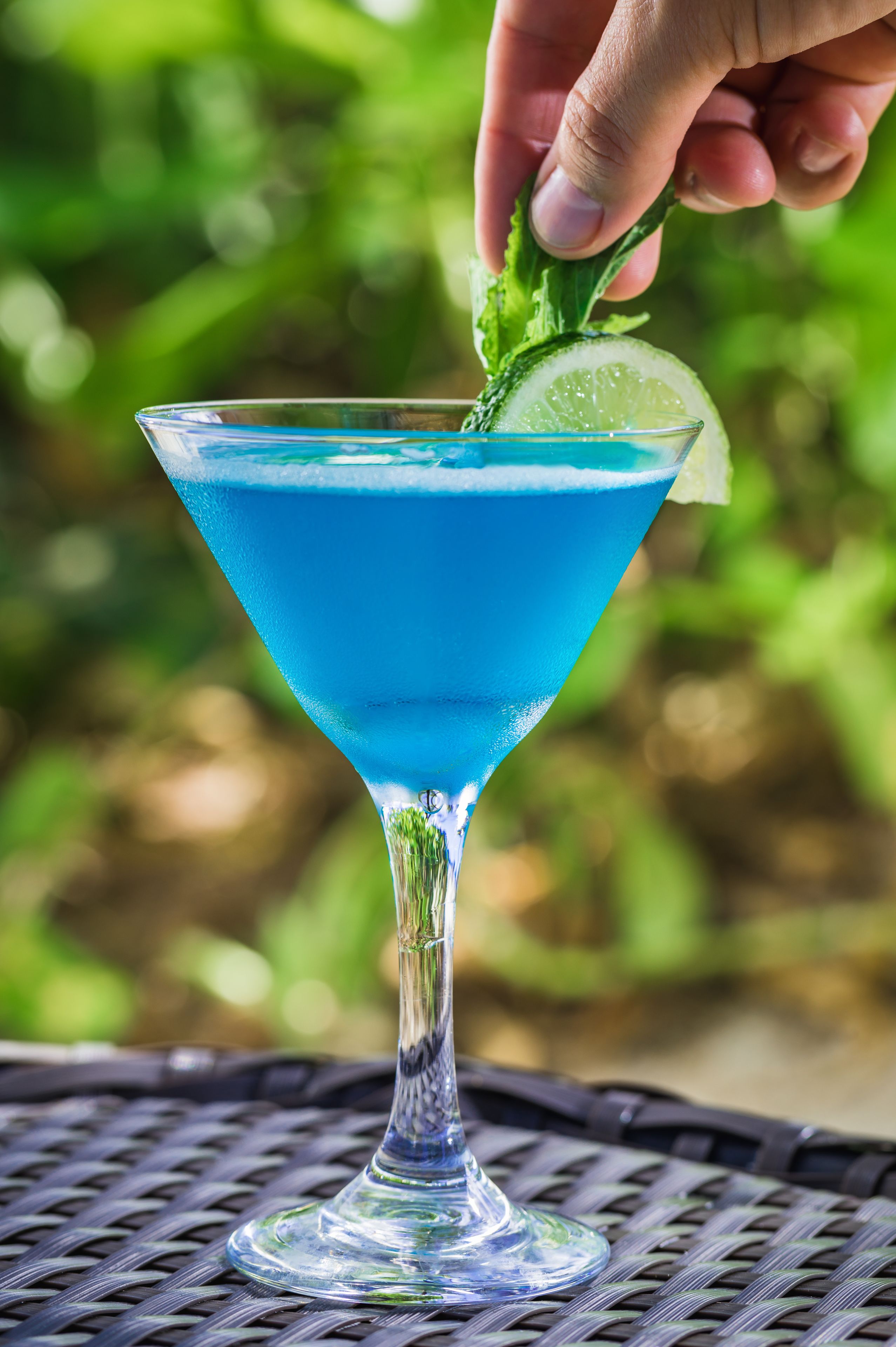 A blue cocktail in a martini glass with a lime wedge on the rim, placed on a woven table.